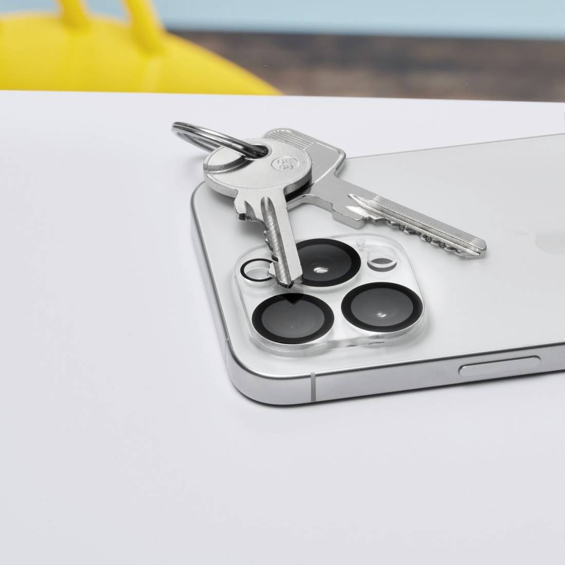 Ein Smartphone liegt flach auf einem Tisch, darauf liegen Schlüssel mit einem silbernen Schlüsselanhänger. Links im Hintergrund unscharf ein gelber Stuhl.