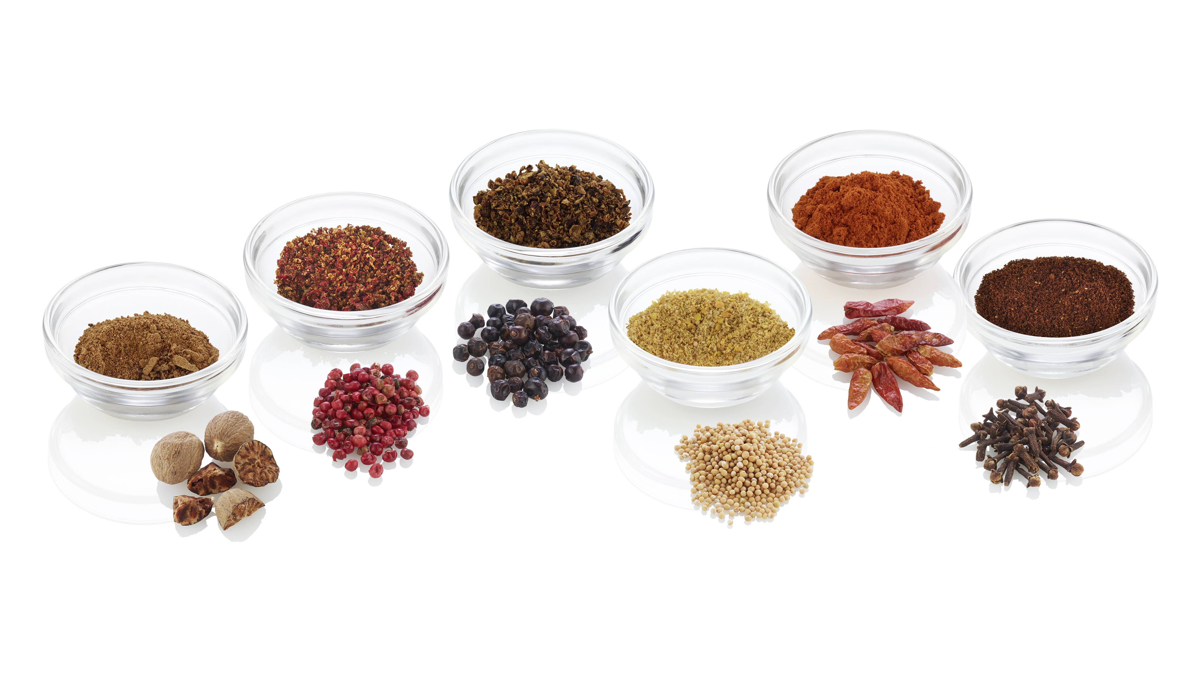 Verschiedene Gewürze in Glasschalen, darunter Paprika, Pfefferkörner und Nelken, auf weißem Hintergrund arrangiert.