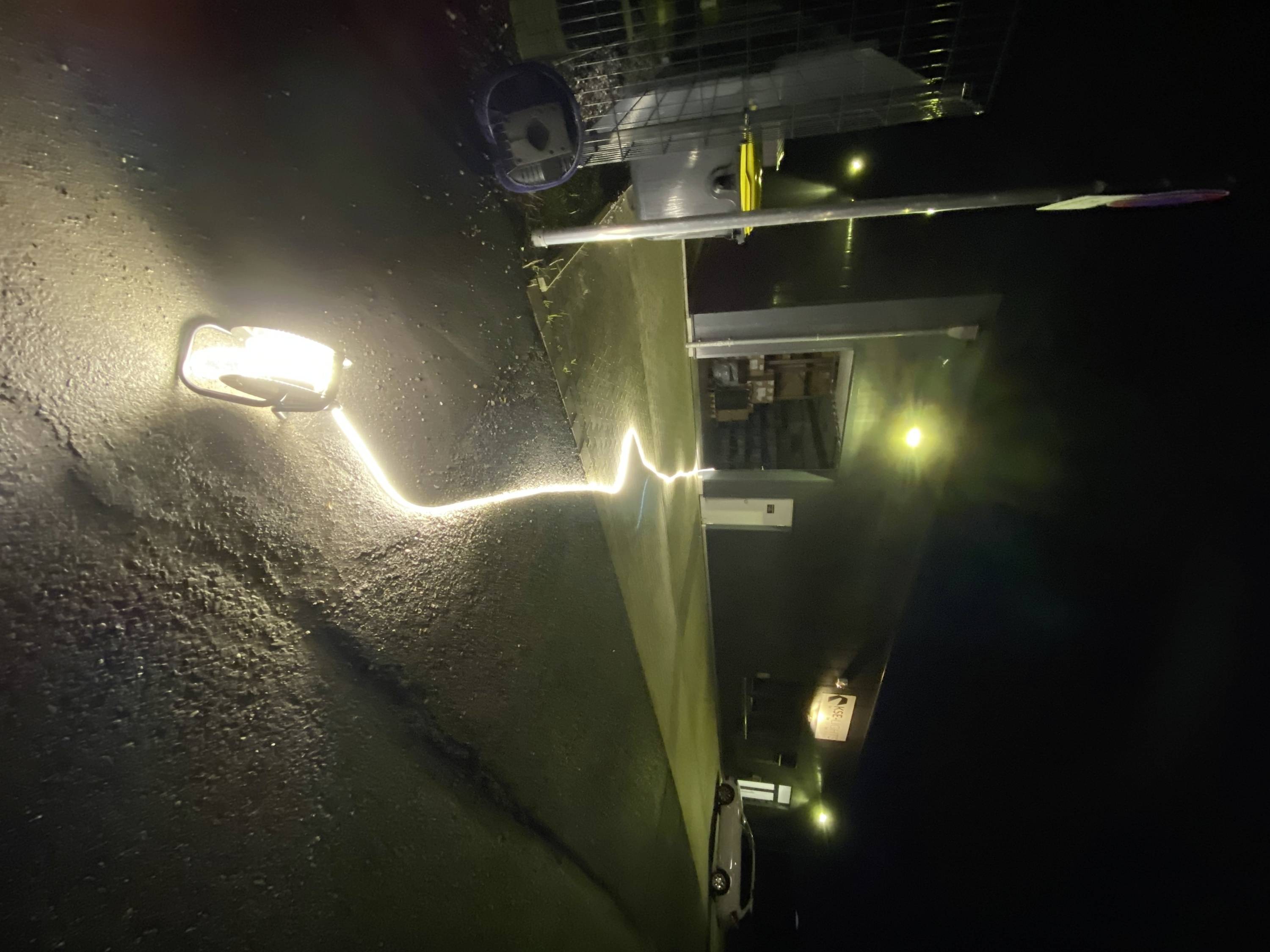 'Nachtaufnahme einer beleuchteten Straße vor einem Industriegebäude. Ein helles Licht markiert einen beleuchteten Weg zum Gebäude.'