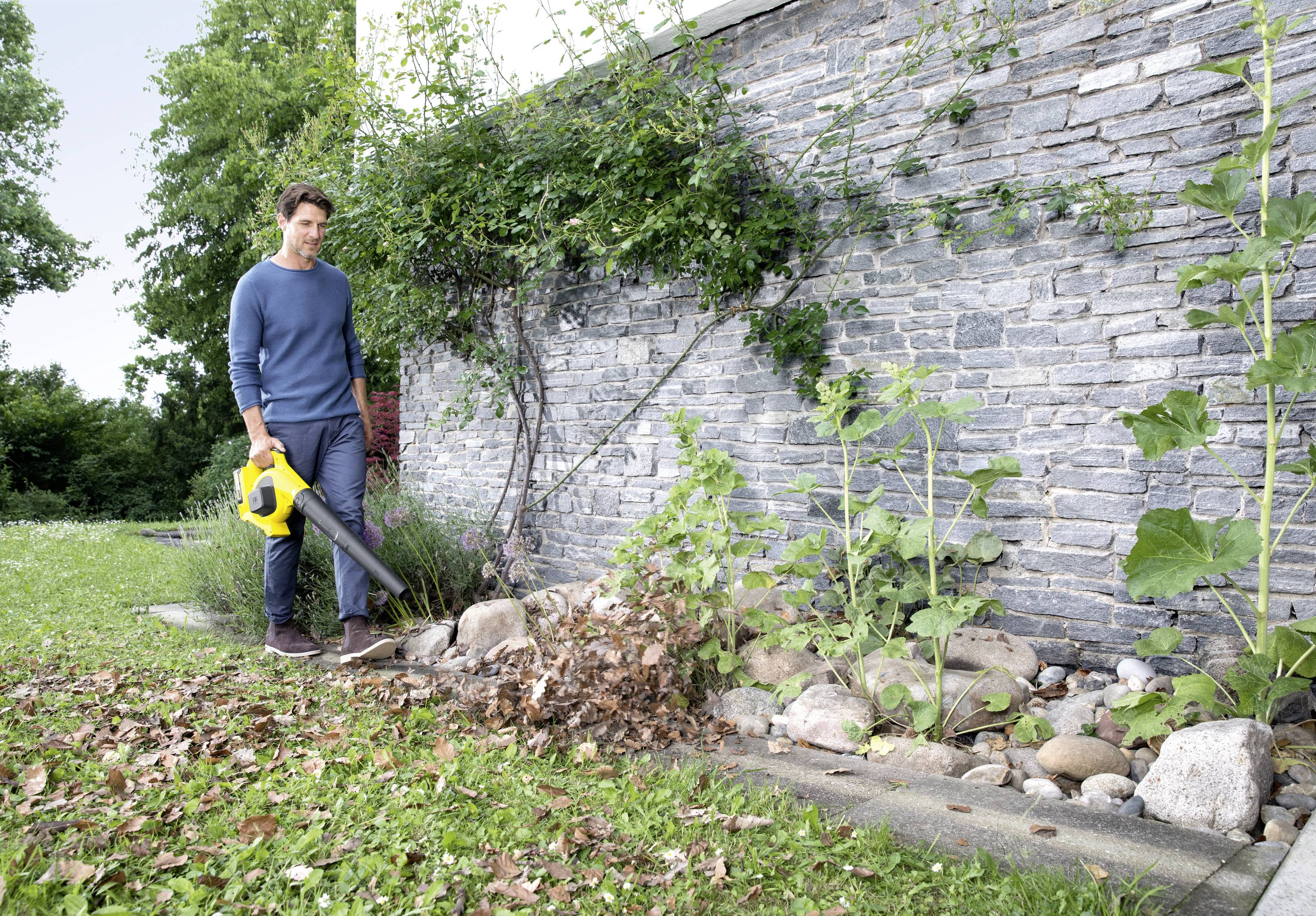 Eine Person benutzt einen Laubbläser, um Laub im Garten entlang einer Steinmauer zu entfernen. Im Hintergrund sind Bäume sichtbar.