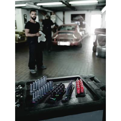 Ein Mechaniker steht in einer Werkstatt. Im Vordergrund sind zahlreiche Steckschlüssel sortiert. Ein rotes Auto ist im Hintergrund sichtbar.
