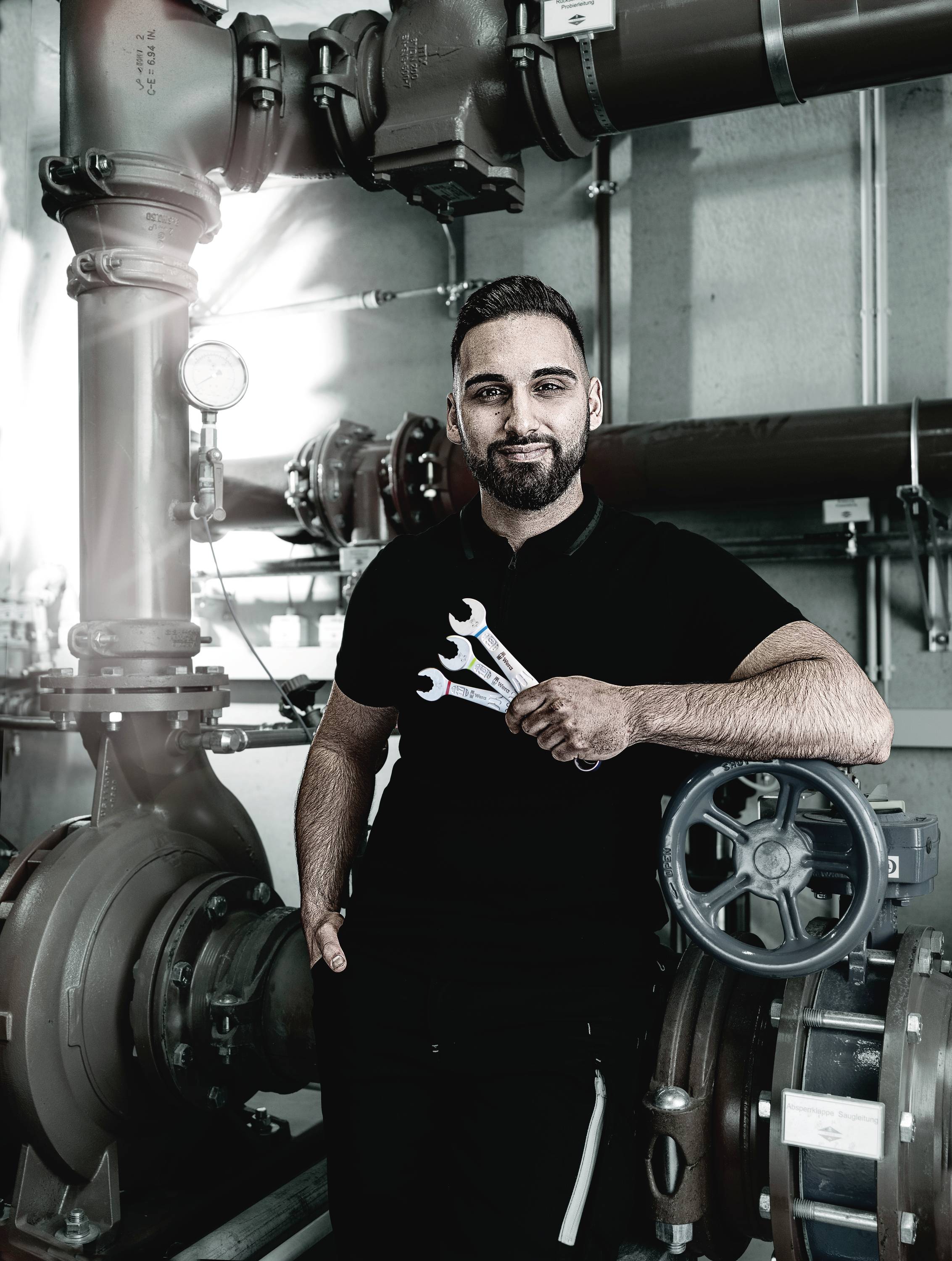 Ein Mann in Arbeitskleidung steht in einer industriellen Anlage. Er hält drei Schraubenschlüssel in der Hand und schaut in die Kamera.