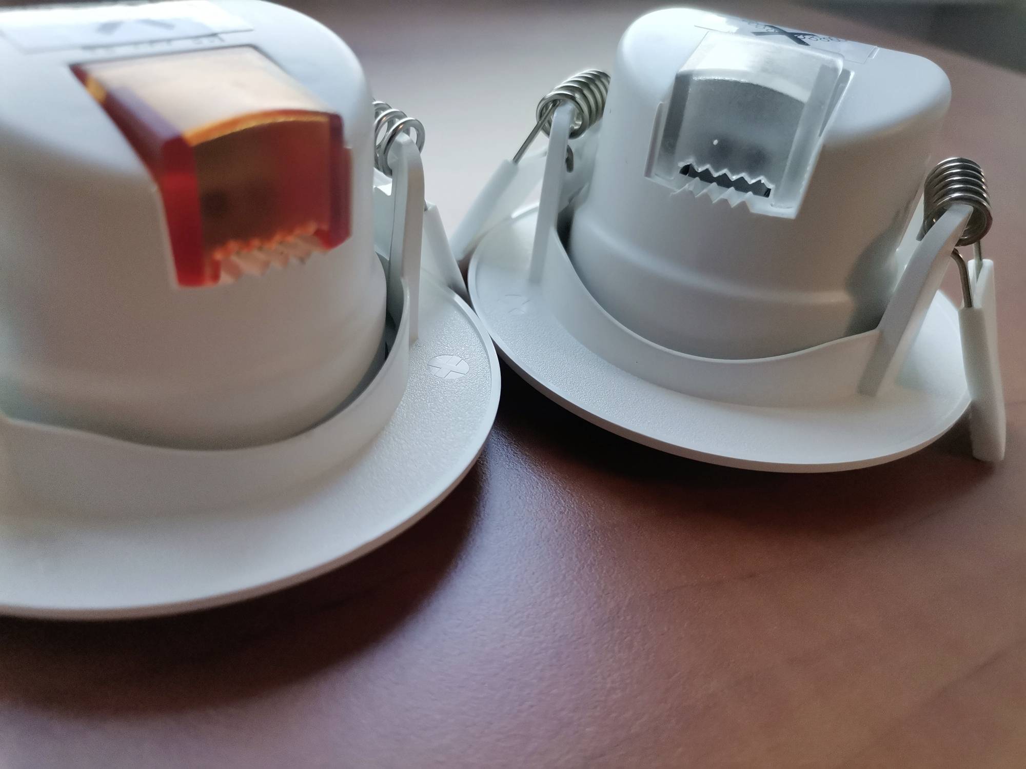 Zwei weiße LED-Deckeneinbauleuchten mit Federn auf einer braunen Oberfläche, eine mit rot getöntem Abdeckung, die andere mit klarem.