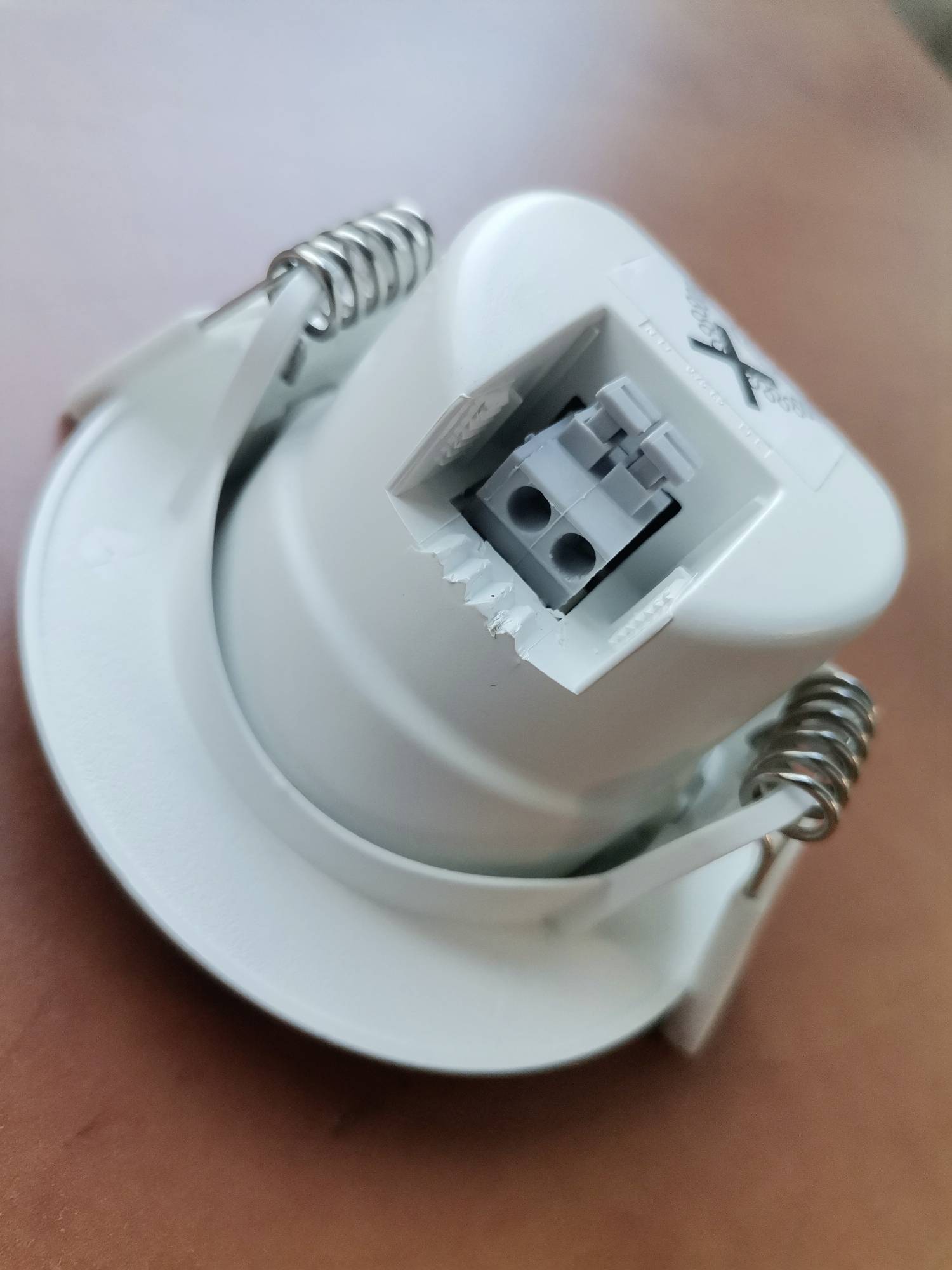 Ein weißer Deckeneinbausockel mit zwei Metallklammern und einer zentralen Anschlussklemme liegt auf einem braunen Tisch.