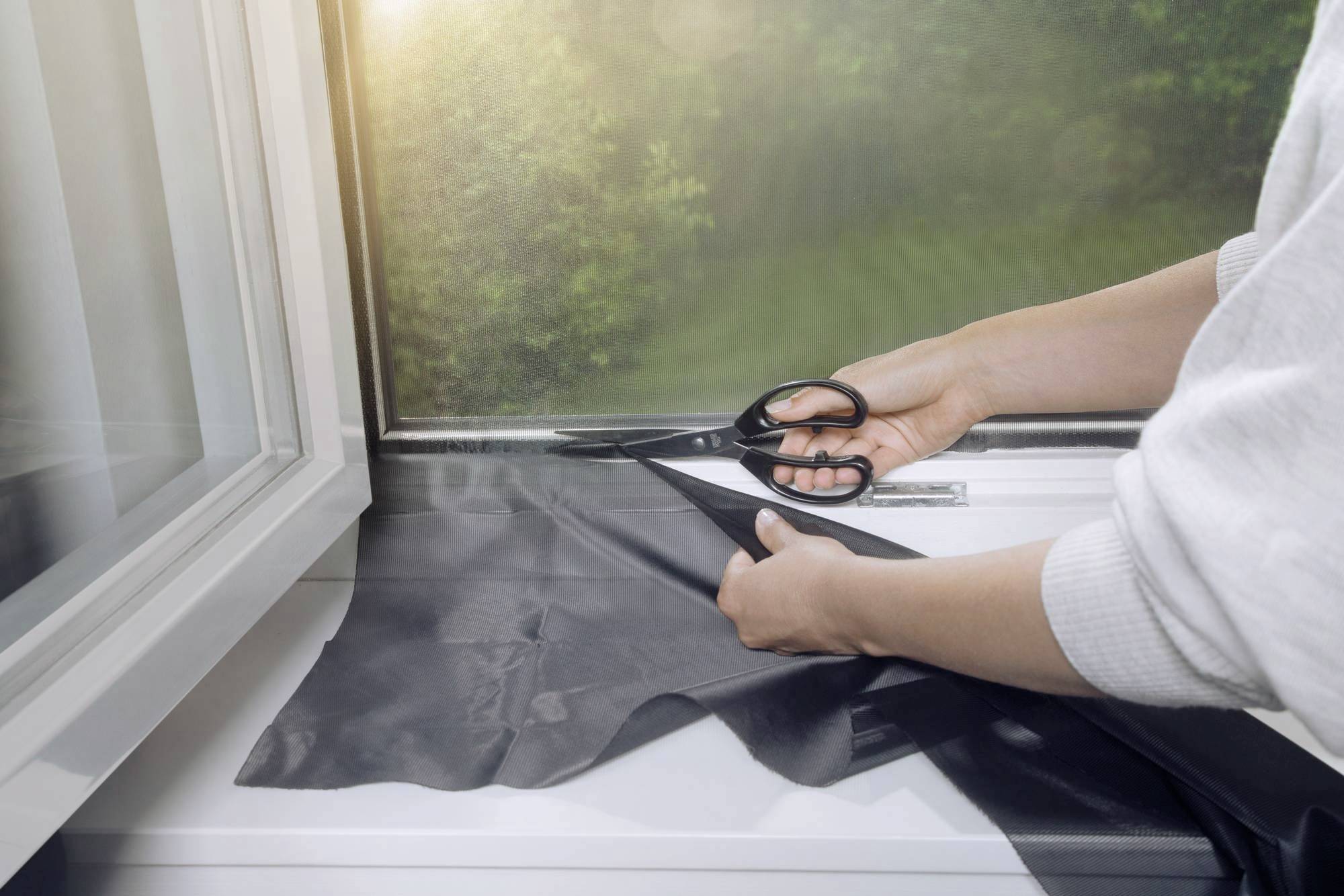 Eine Person schneidet schwarze Folie an einem geöffneten Fenster. Im Hintergrund ist ein grüner Garten zu sehen.