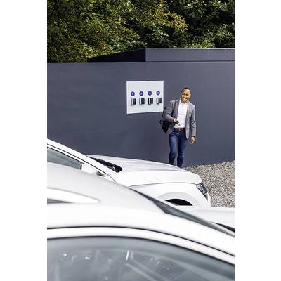 Eine Person in formeller Kleidung lächelt, während sie an einer Reihe von Ladestationen für Elektroautos vorbeigeht. Im Vordergrund sind Autos zu sehen.