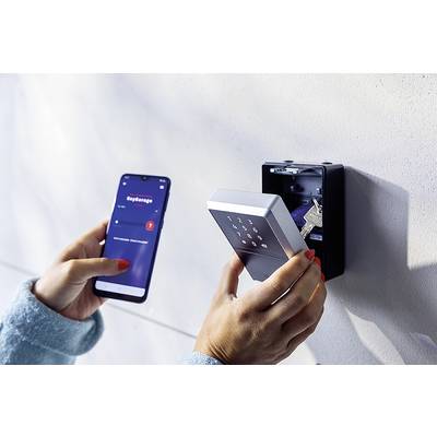 Eine Person hält ein Smartphone mit einer geöffneten App in der linken Hand und einen elektronischen Schlüsselsafe in der rechten.