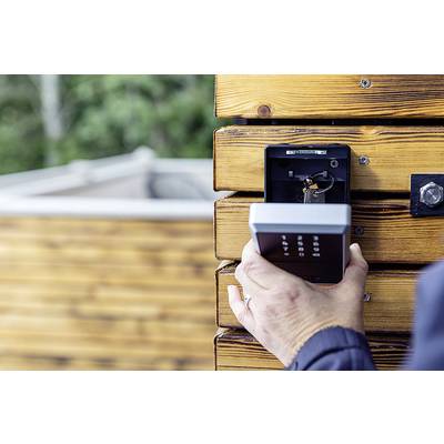 Eine Person hält ein Zahlenschlossgerät, das an einer Holzwand montiert ist. Im Hintergrund ist ein unscharfer, grasbewachsener Bereich zu sehen.