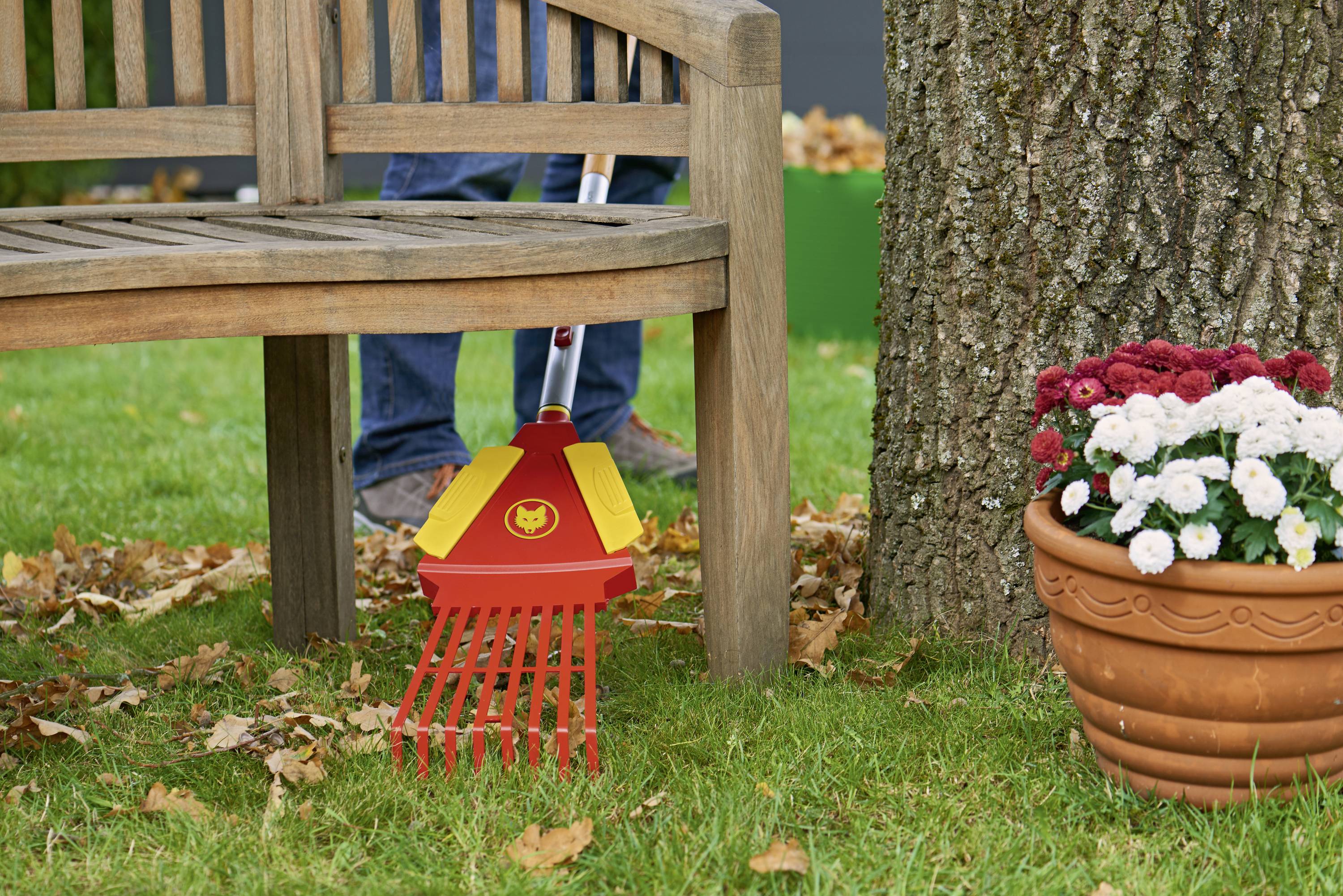 Ein roter Laubrechen neben einer Holzbank und einem Baum, mit Blumen in einem Topf daneben. Blätter liegen verstreut auf dem Rasen.