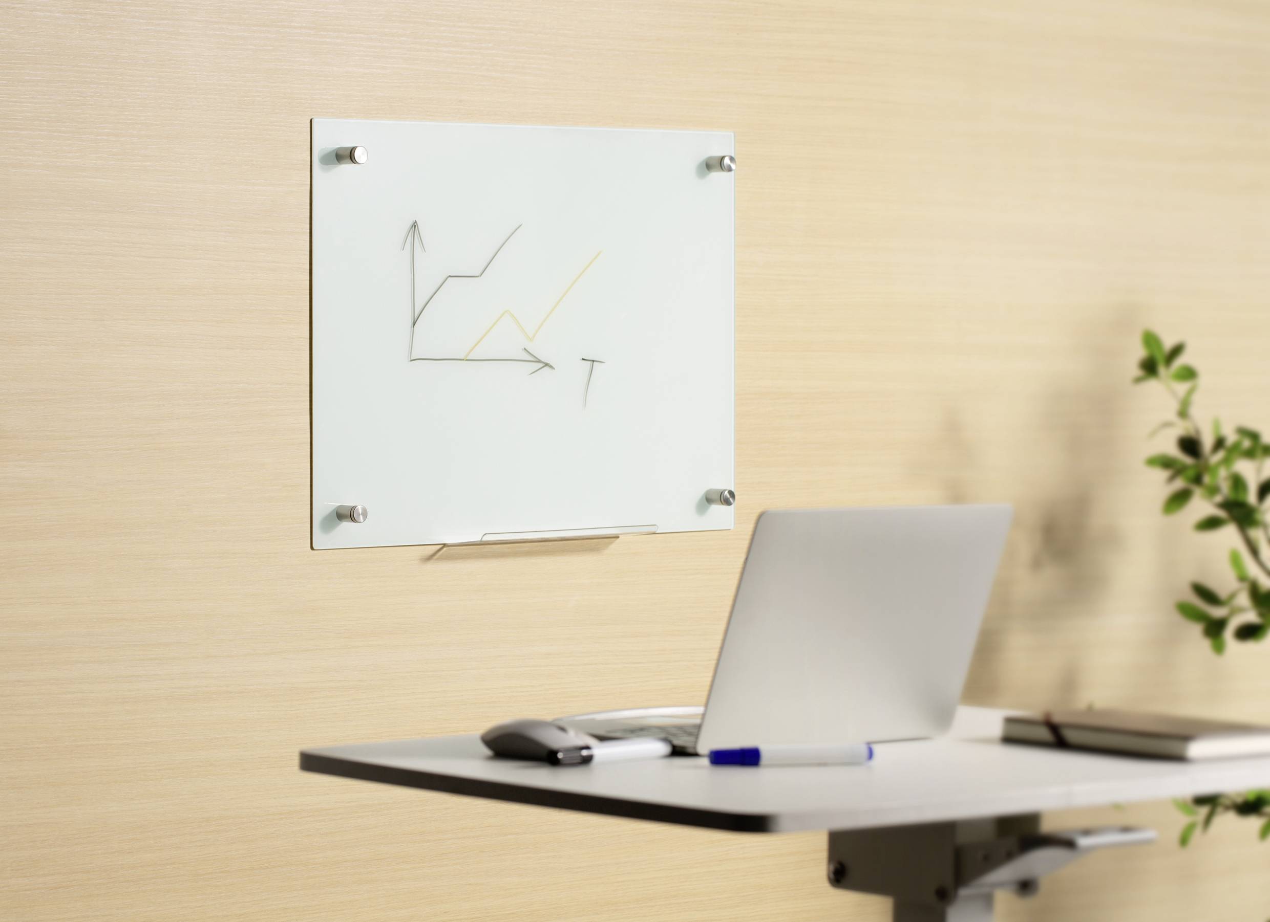 Laptop auf einem Schreibtisch, daneben ein Diagramm mit ansteigenden Linien an der Wand. Eine Pflanze ist im Hintergrund sichtbar.