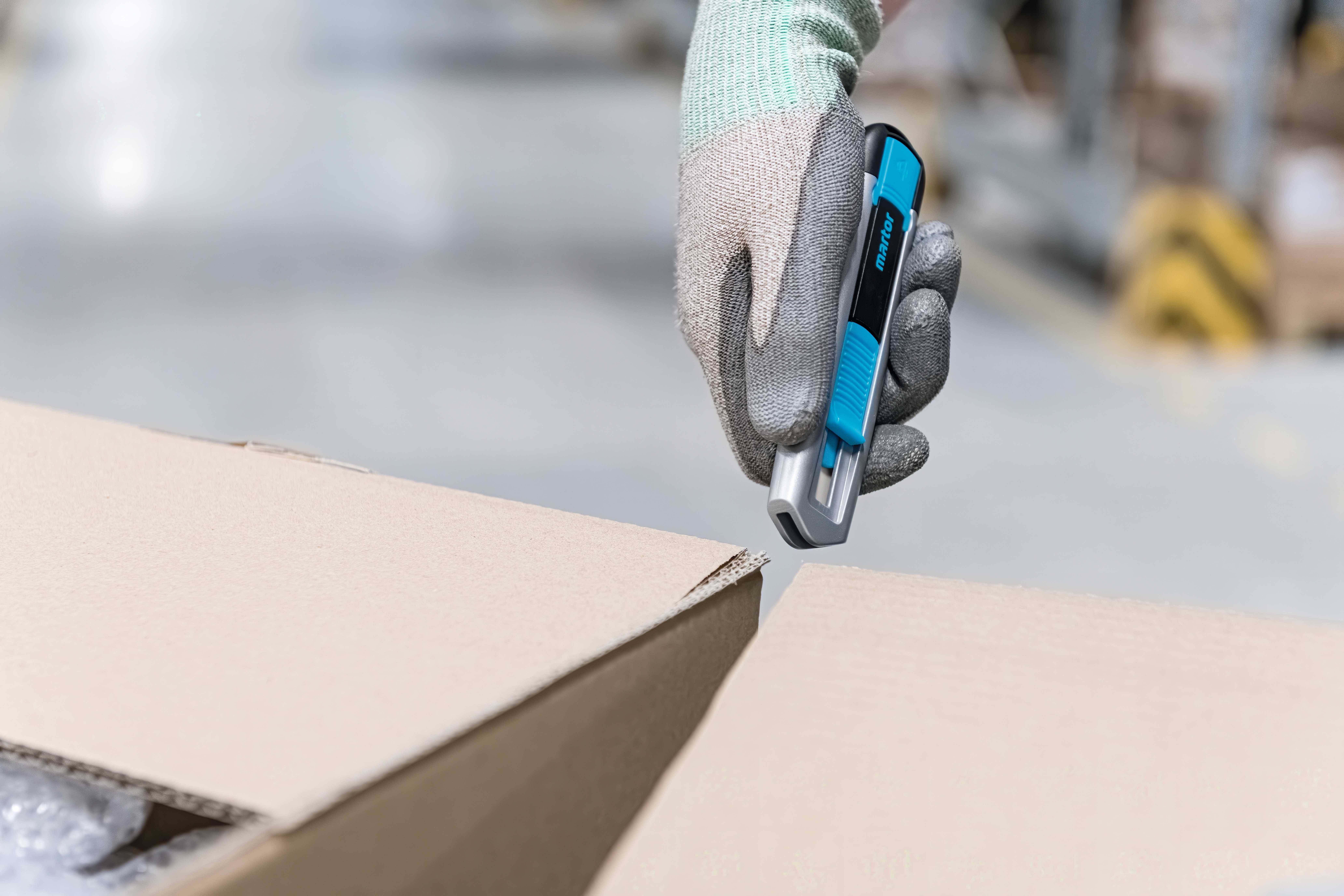 Eine Person mit grauen Handschuhen hält einen blauen Cutter, um einen Karton in einem Lager zu öffnen.