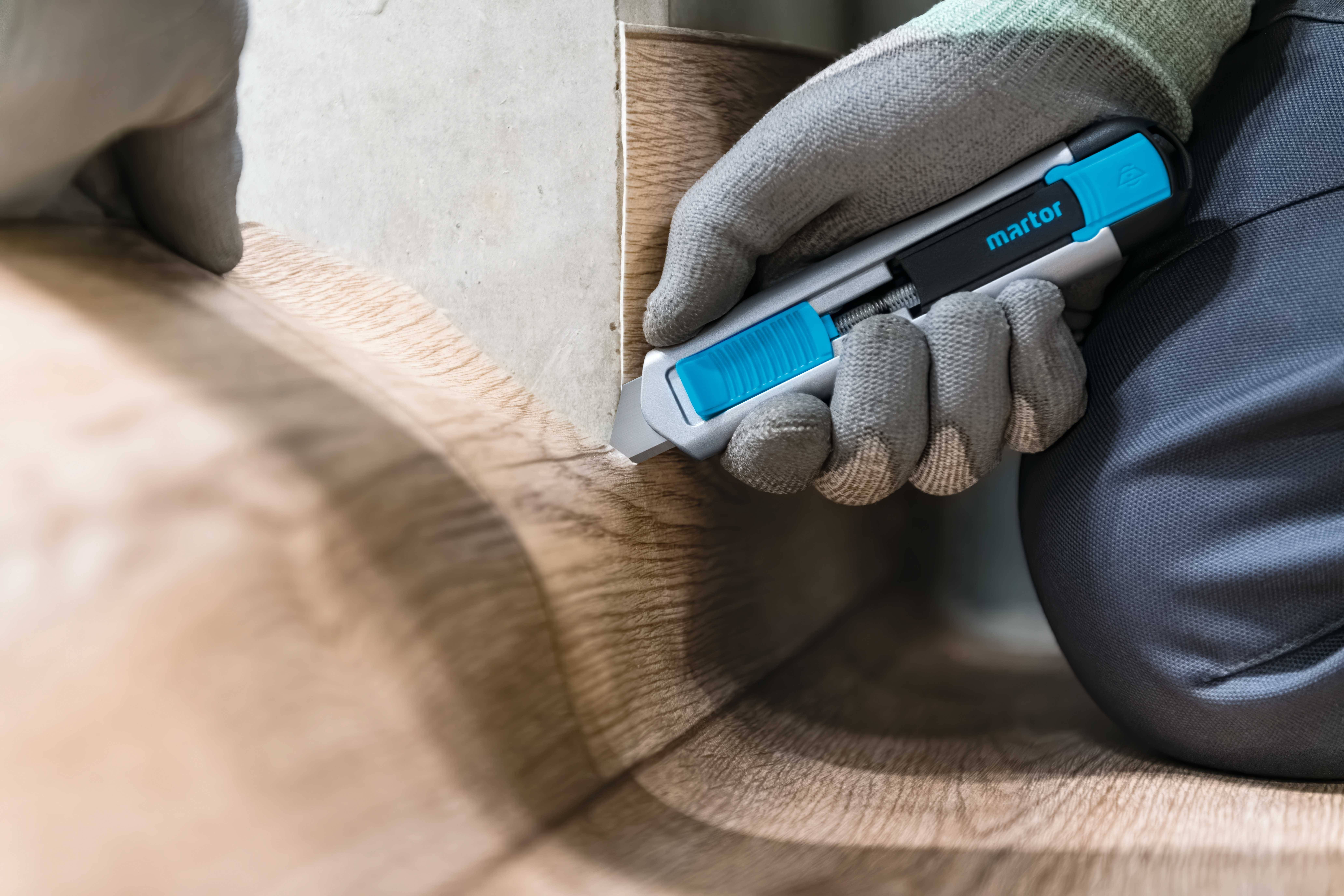Eine Person schneidet mit einem blauen Cutter-Messer und grauen Handschuhen einen Bodenbelag präzise an einer Ecke zurecht.
