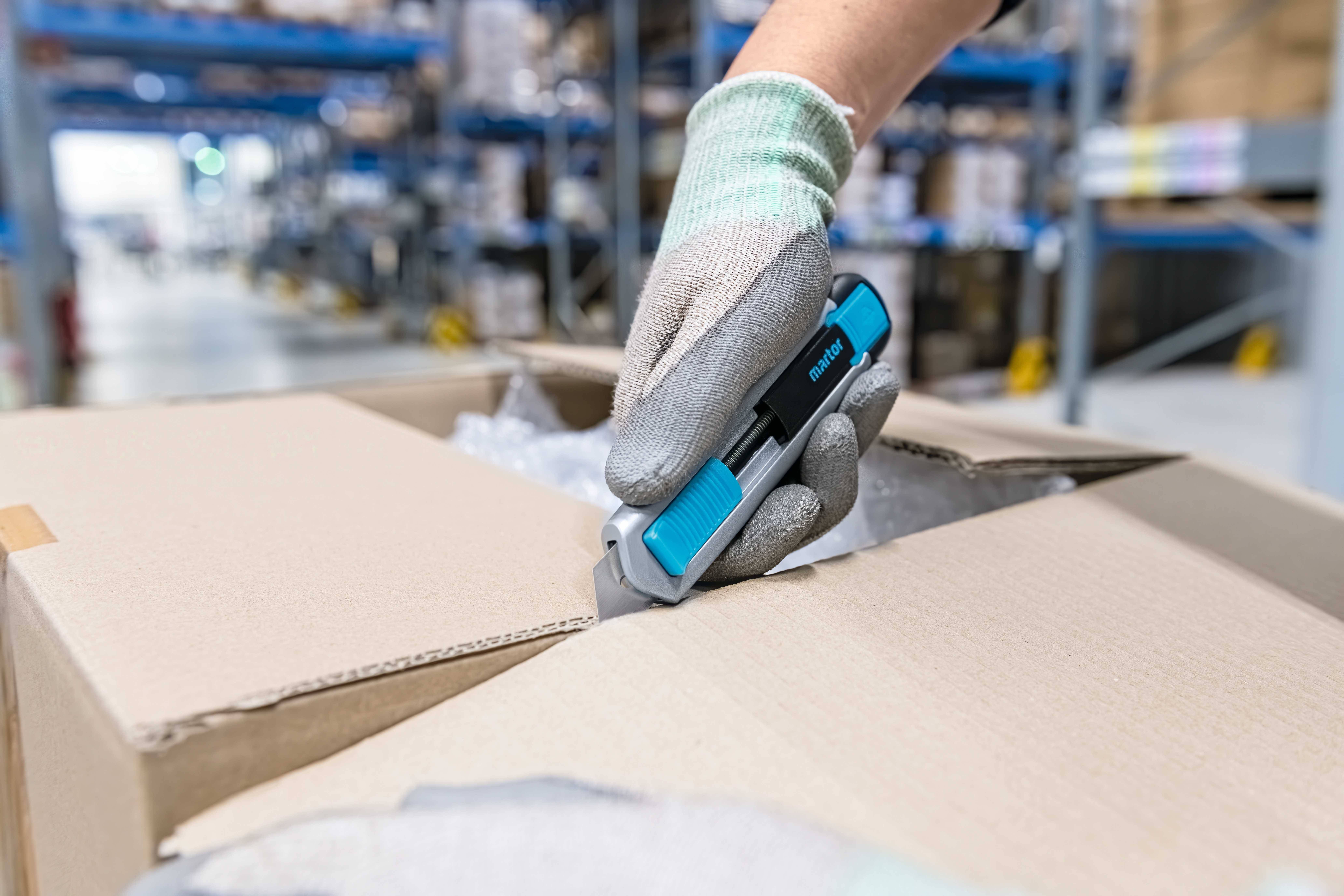 Eine Hand mit Handschuh öffnet mit einem blauen Sicherheitsmesser einen Pappkarton in einem Lagerhaus. Im Hintergrund sind Regale zu sehen.