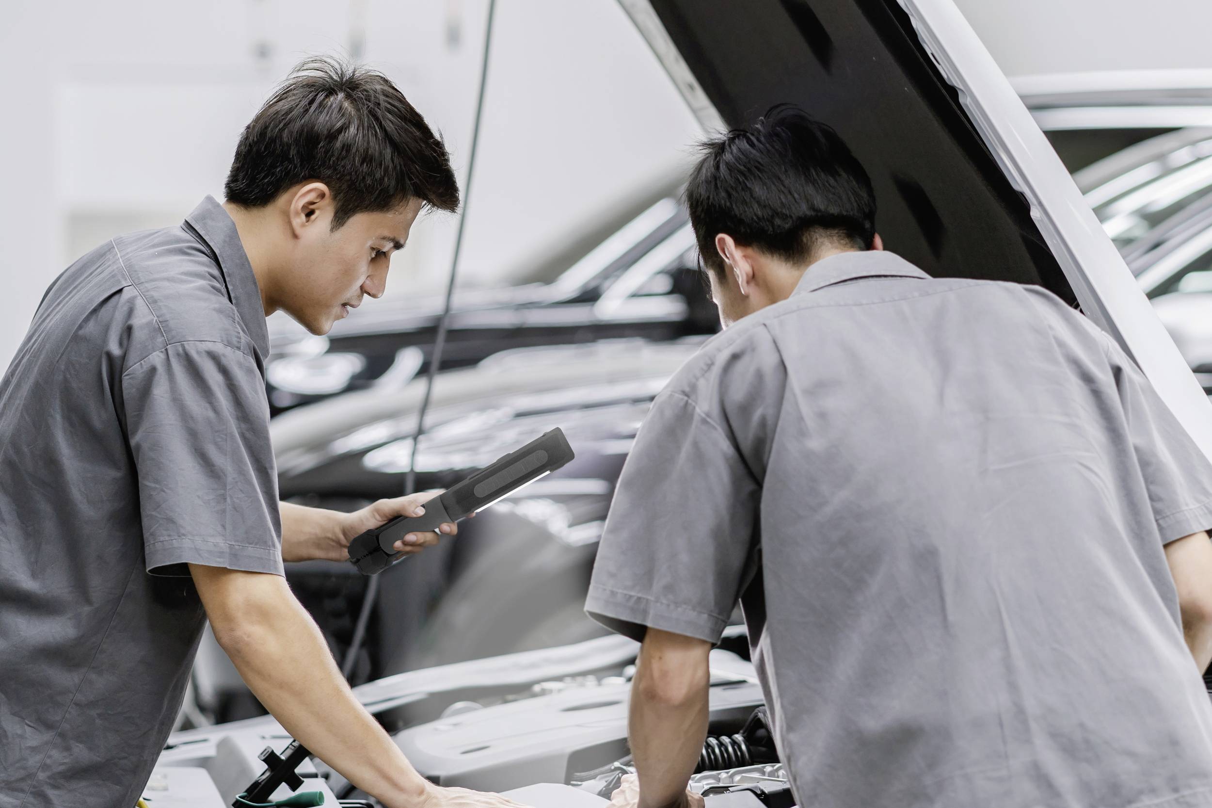 Zwei Mechaniker untersuchen den Motor eines Autos in einer Werkstatt. Einer hält ein digitales Diagnosegerät in der Hand.