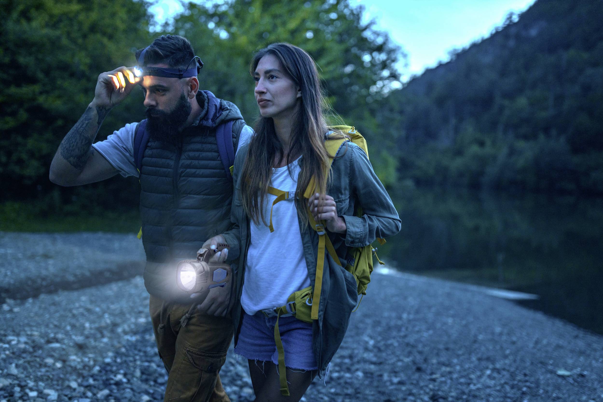 Ein Mann und eine Frau wandern am Flussufer entlang eines bewaldeten Gebiets bei Dämmerung. Beide tragen Rucksäcke und Taschenlampen.