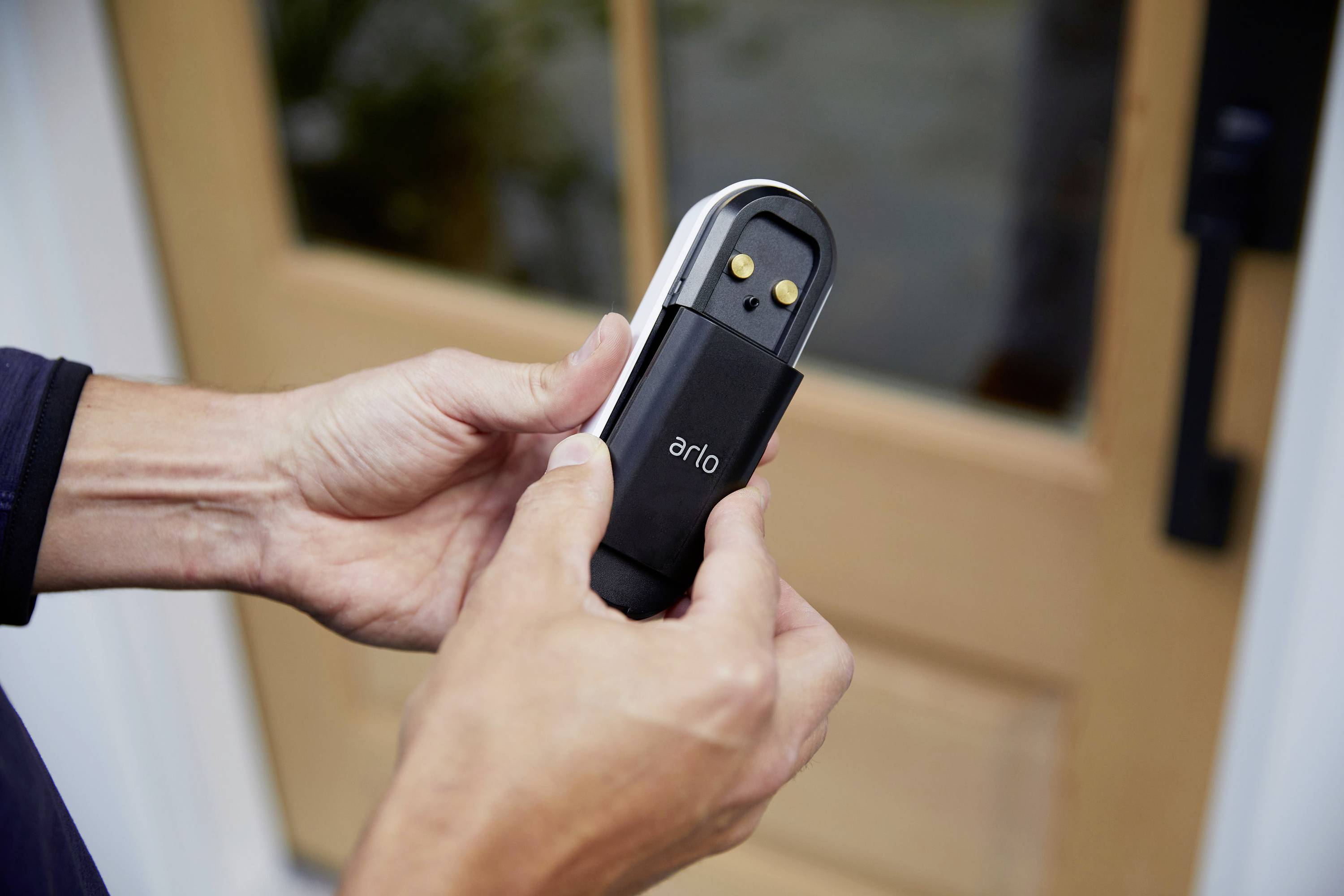Eine Person hält eine Arlo-Türklingel vor einer geschlossenen Holztür. Die Türklingel hat zwei goldene Kontaktpunkte.