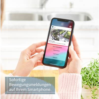 Eine Hand hält ein Smartphone mit einer Benachrichtigung zur Bewegungserkennung. Text: 'Sofortige Bewegungsmeldungen auf Ihrem Smartphone.' Pflanzen im Vordergrund.