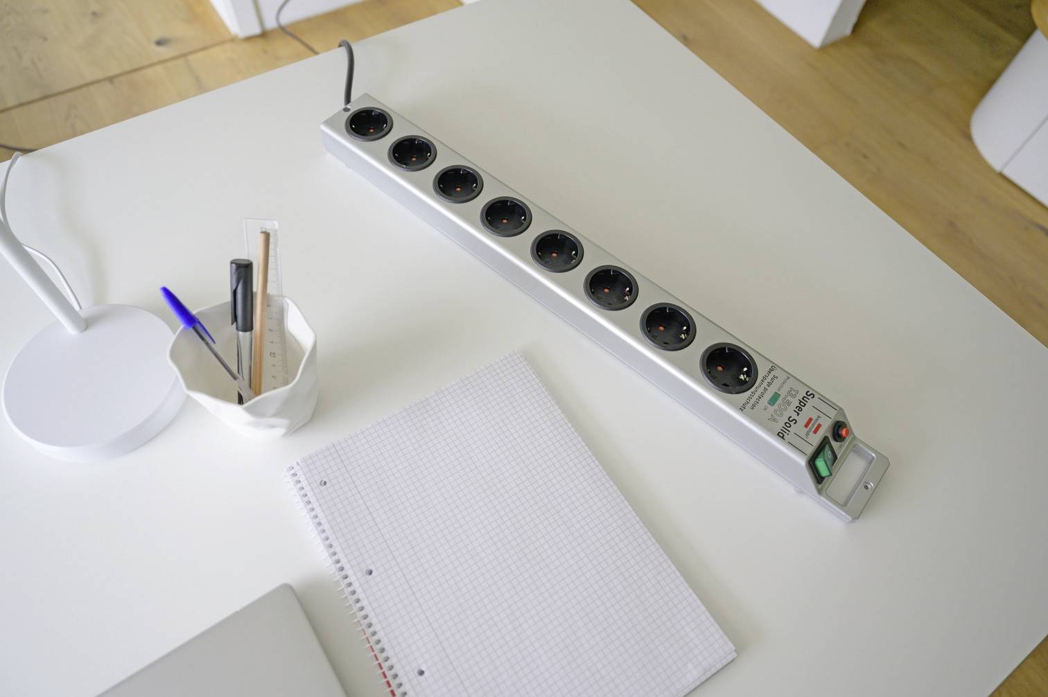 Mehrfachsteckdose auf einem weißen Tisch neben einem Block Papier und einem Behälter mit Stiften, in einem Büro- oder Arbeitsumfeld.