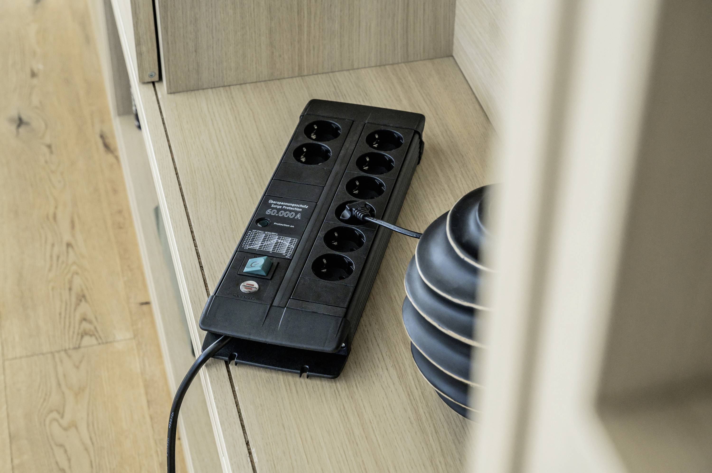 Eine schwarze Mehrfachsteckdose mit Schalter und LED-Anzeige liegt auf einem Holzregal. Ein Kabel ist eingesteckt.