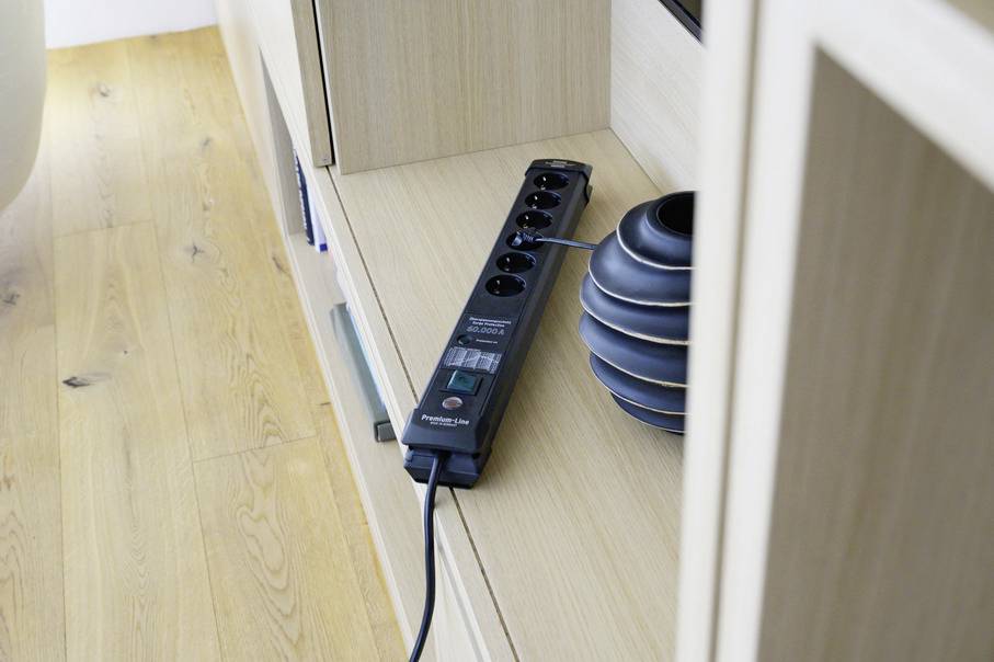 Eine schwarze Steckdosenleiste liegt auf einem hellen Holzmöbel. Daneben steht ein geriffeltes, graues Gefäß.