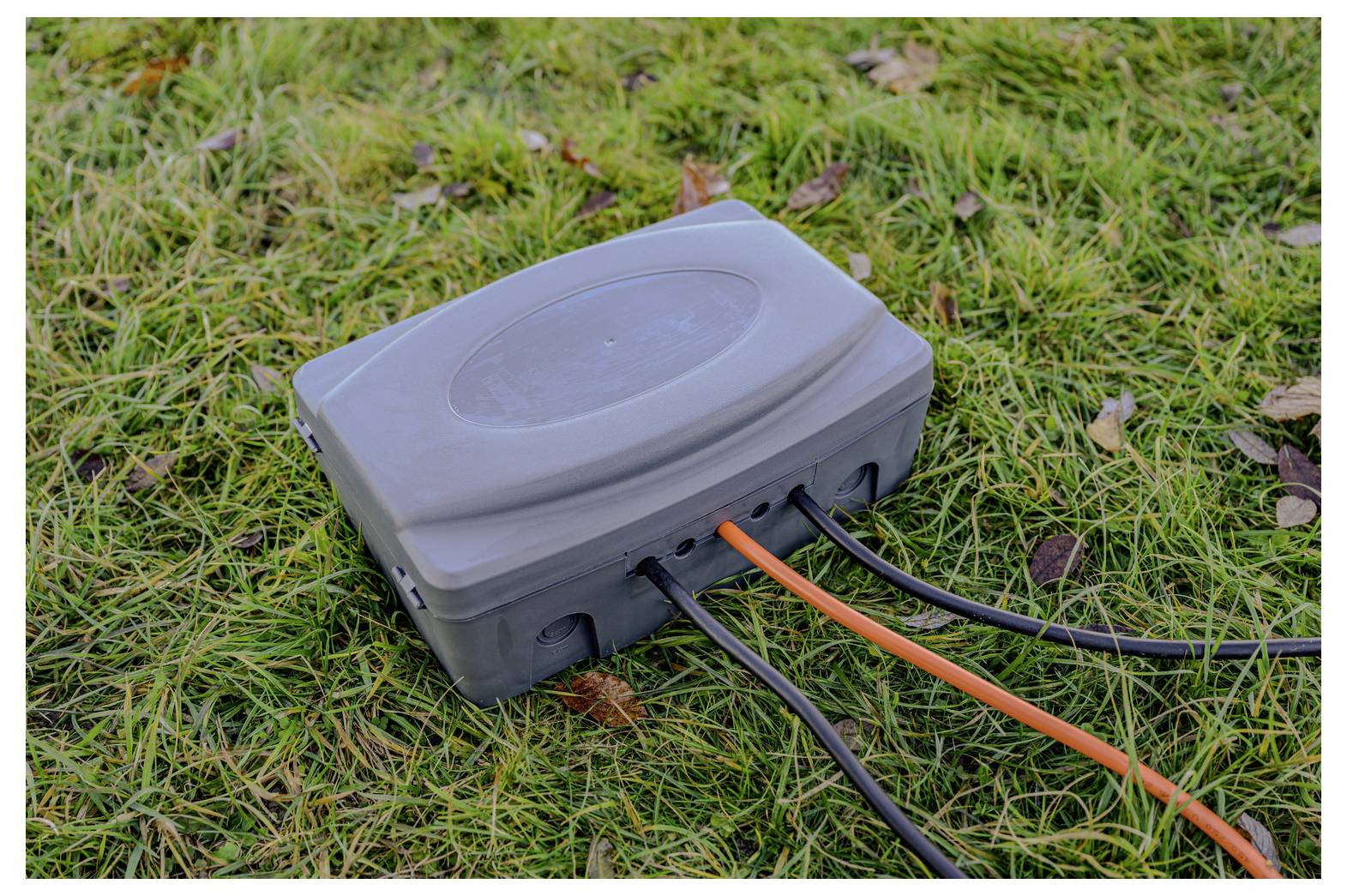 Graue Außenelektrik-Schaltkasten auf Gras mit mehreren schwarzen und orangefarbenen Kabeln verbunden.