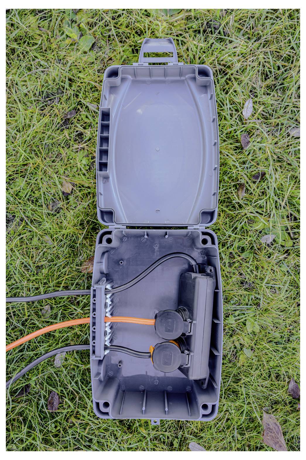 Offene schwarze Kabelverbindungskammer auf Gras mit angebrachtem Deckel, zeigt verbundene orange und schwarze Kabel sowie interne Verkabelung.