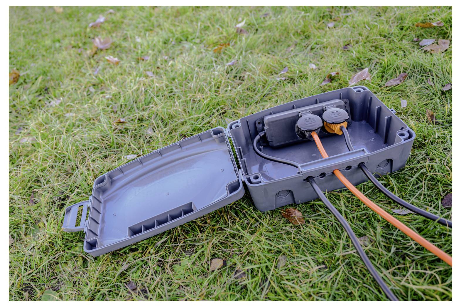Offene wasserdichte elektrische Anschlussdose auf Gras mit orangefarbenen und schwarzen Kabeln, die innen verbunden sind.