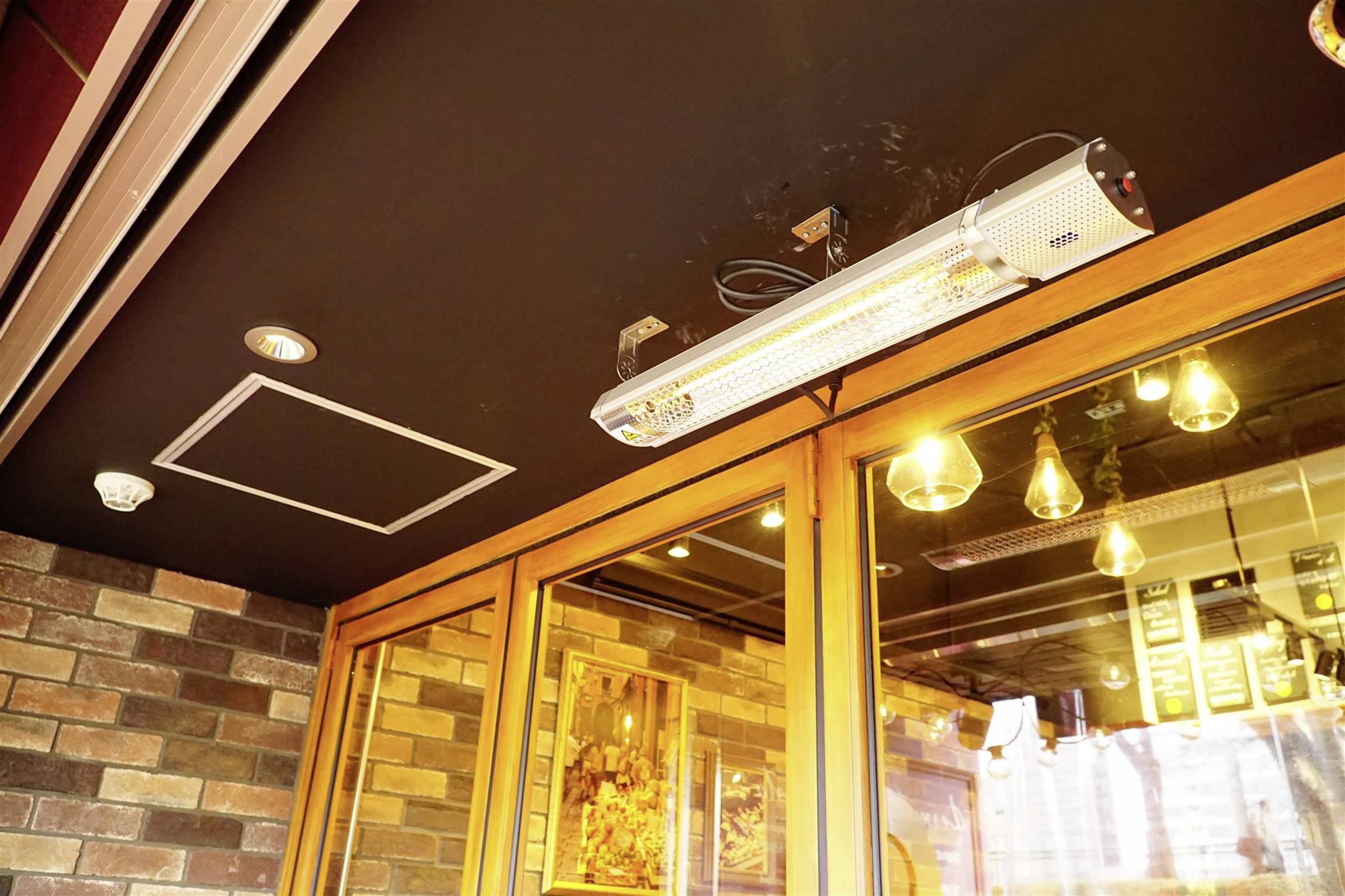 Heizstrahler hängt unter der Decke eines Cafés mit Backsteinwänden. Reflektierende Fenster zeigen Lichter und Innenraum.