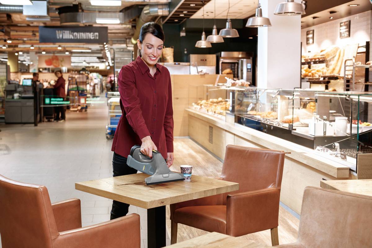 Eine Frau in rotem Hemd reinigt mit einem kleinen Staubsauger einen Holztisch in einem Café oder einer Bäckerei. Im Hintergrund Regale mit Gebäck.