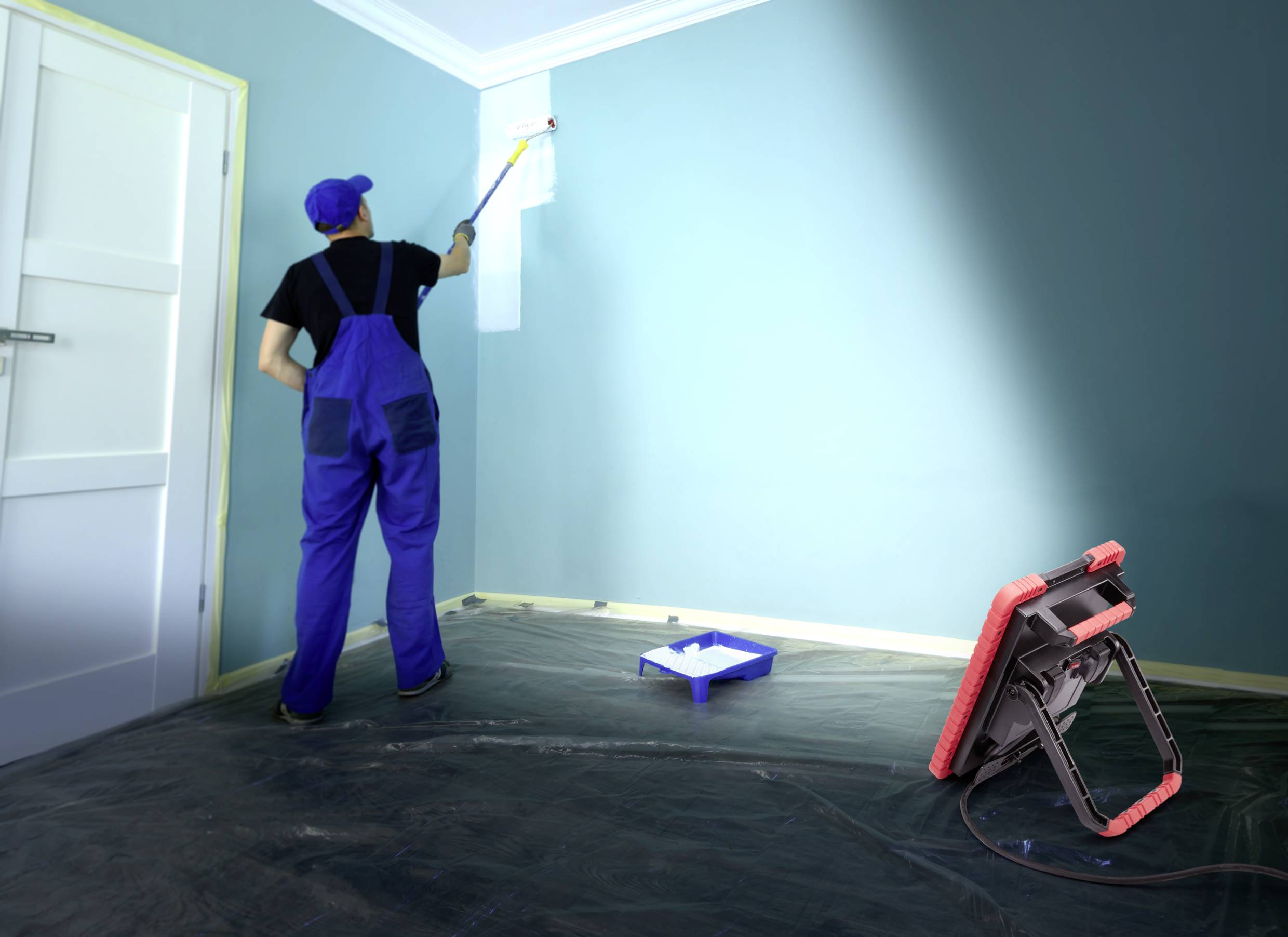 Eine Person in blauer Arbeitskleidung streicht eine Zimmerwand mit weißer Farbe. Eine Abdeckplane schützt den Boden, und eine Lampe leuchtet den Bereich aus.