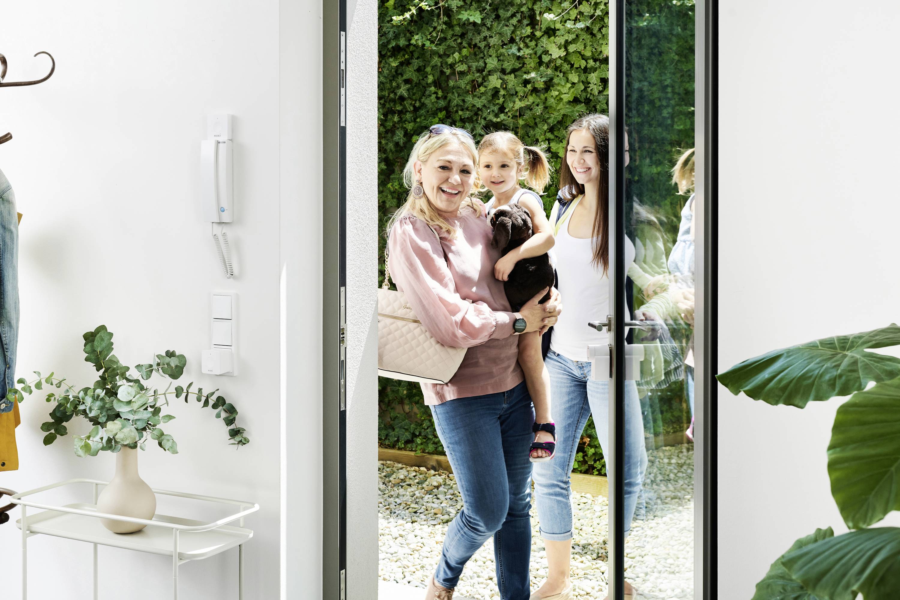 Zwei Frauen, eine hält ein Kind, lächeln und stehen in einer offenen Tür mit Blick nach drinnen. Im Hintergrund sind Pflanzen sichtbar.