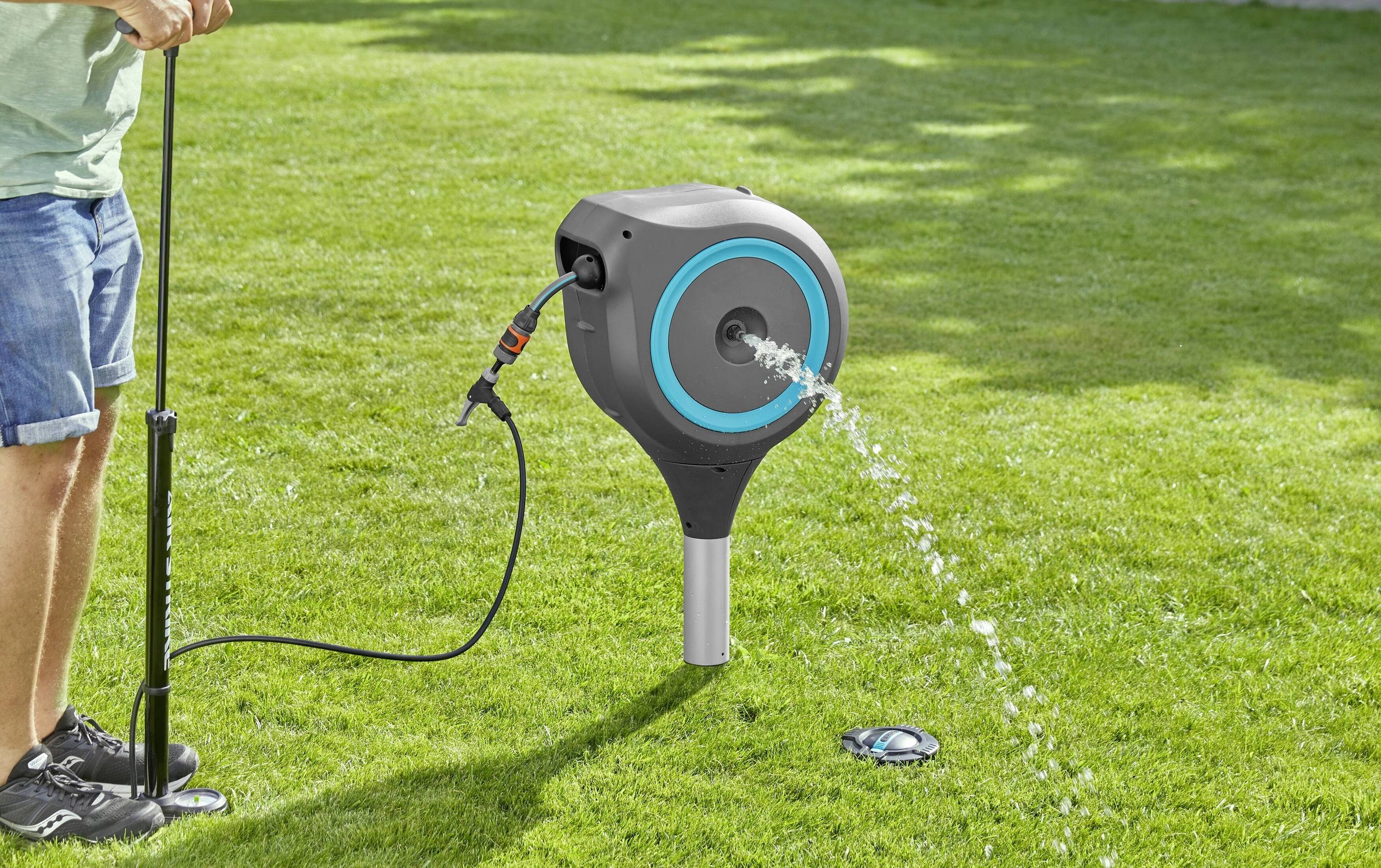 Ein Mensch bedient eine Wasserpumpe auf einem Rasen. Ein Wasserschlauch ist an eine runde, blaue Pumpe angeschlossen und spritzt Wasser heraus.