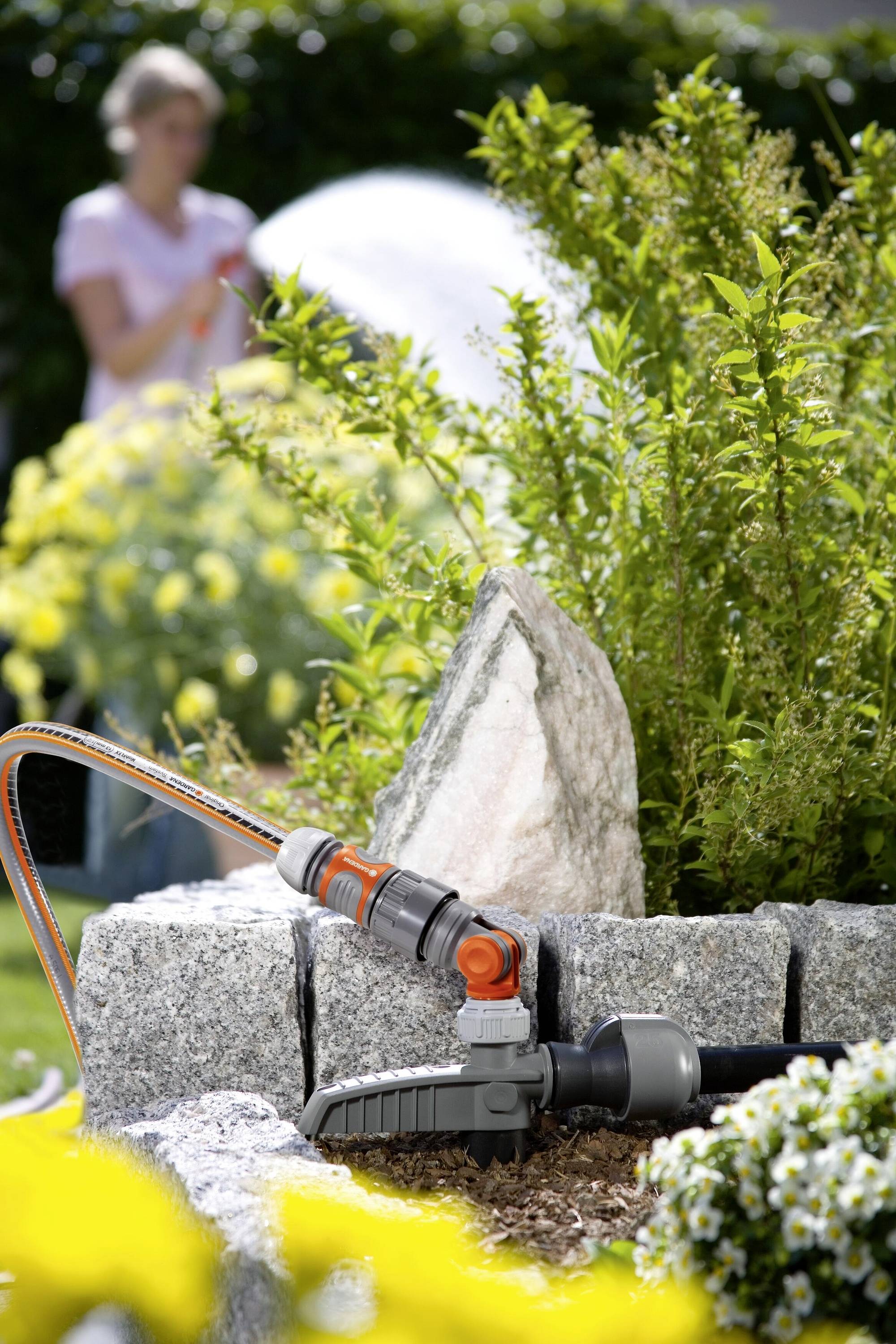Ein Gartenbewässerungssystem mit Schlauch und Sprinkler bewässert Blumenbeete. Eine Person in unscharfem Hintergrund gießt Pflanzen.