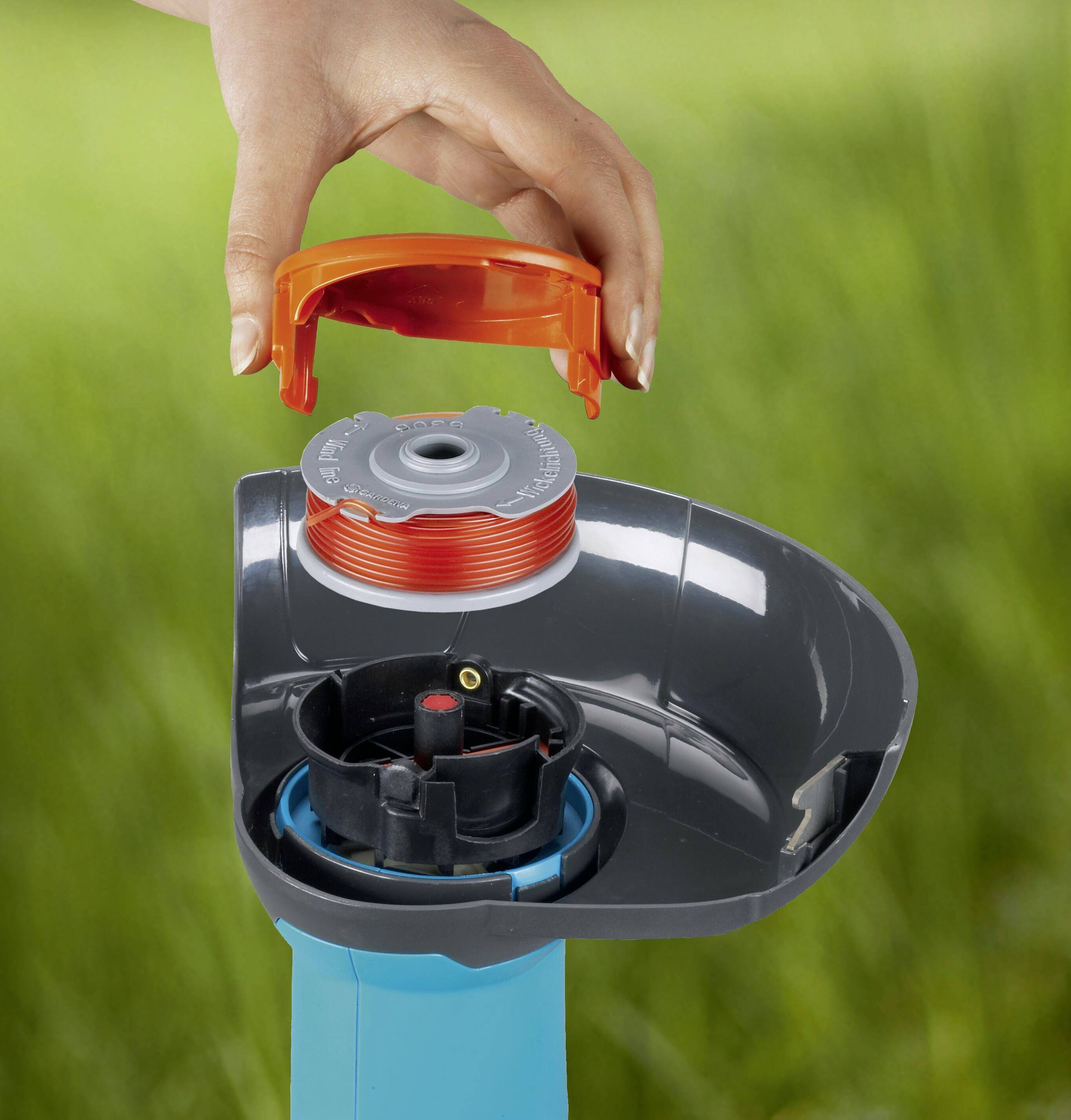 Eine Hand tauscht eine orangene Abdeckung eines blauen Gartengeräts, möglicherweise ein Rasentrimmer, gegen eine Spule aus.