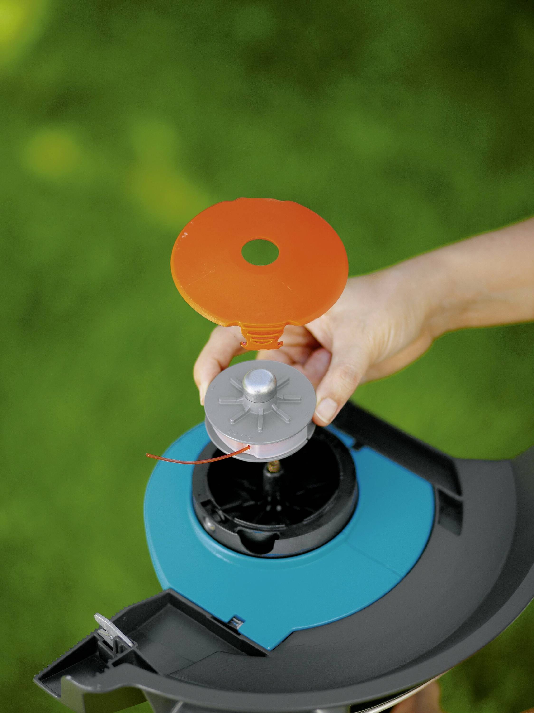 Eine Hand hält einen orangefarbenen Schlüssel über eine grau-blaue Gartenwerkzeugabdeckung. Hintergrund unscharf, grüne Natur.