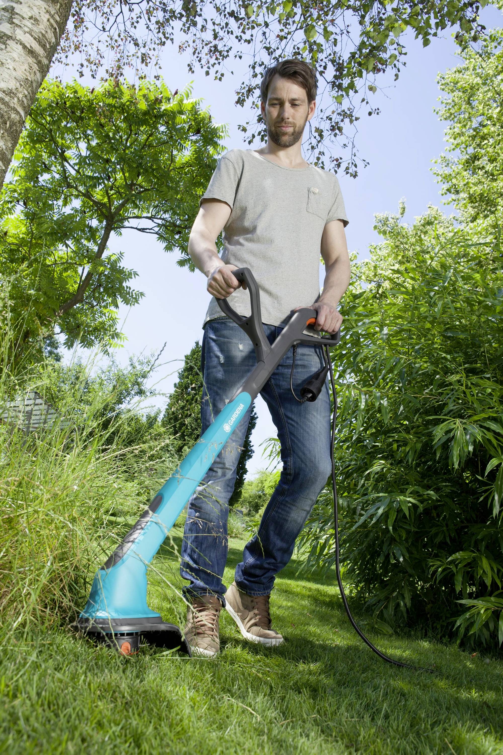 Ein Mann verwendet einen elektrischen Rasentrimmer im Garten. Er trägt lässige Kleidung und arbeitet konzentriert auf einer Rasenfläche.