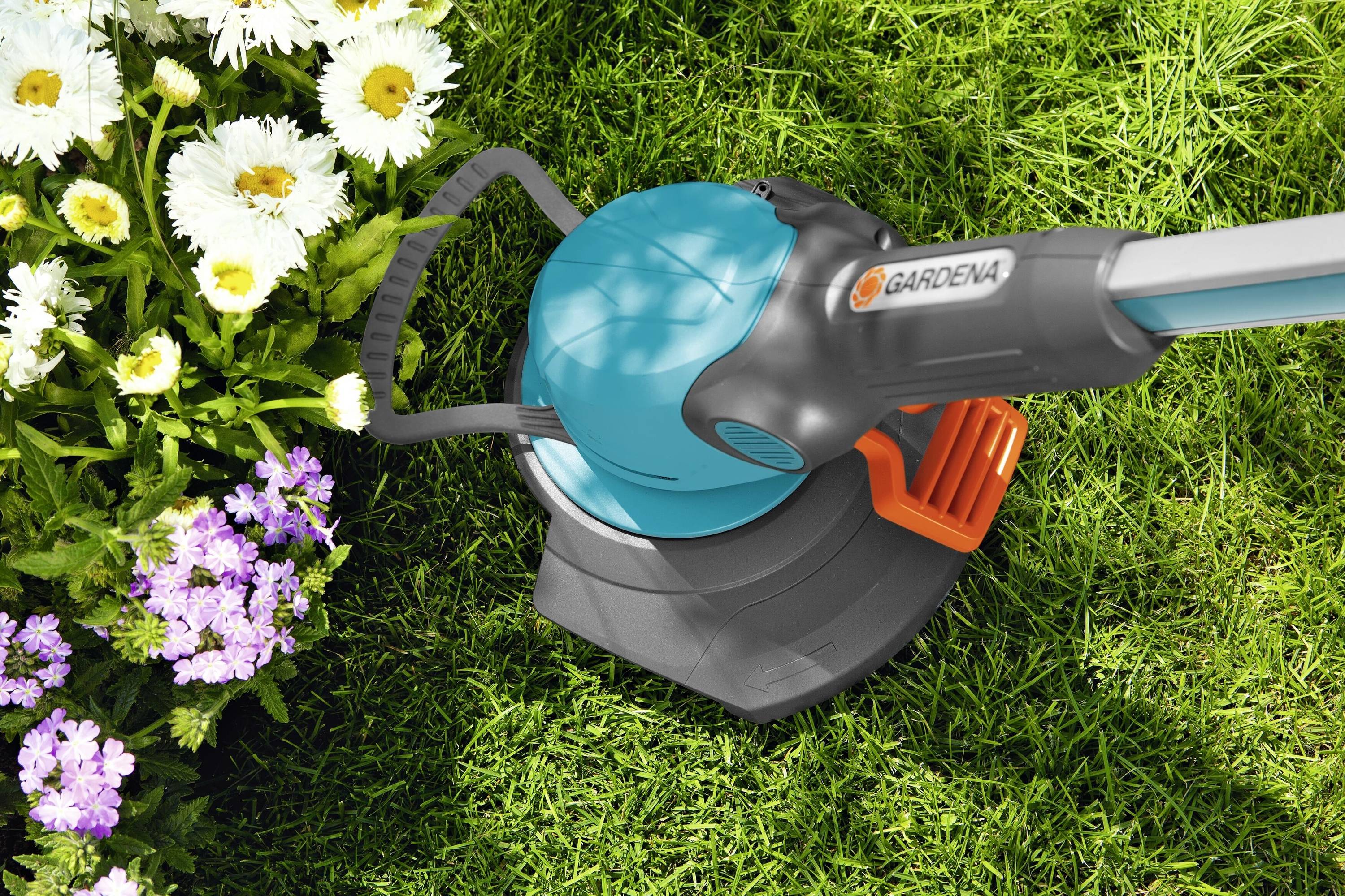 Ein elektrischer Rasentrimmer schneidet Gras an der Kante eines Blumenbeets mit weißen und lila Blüten.