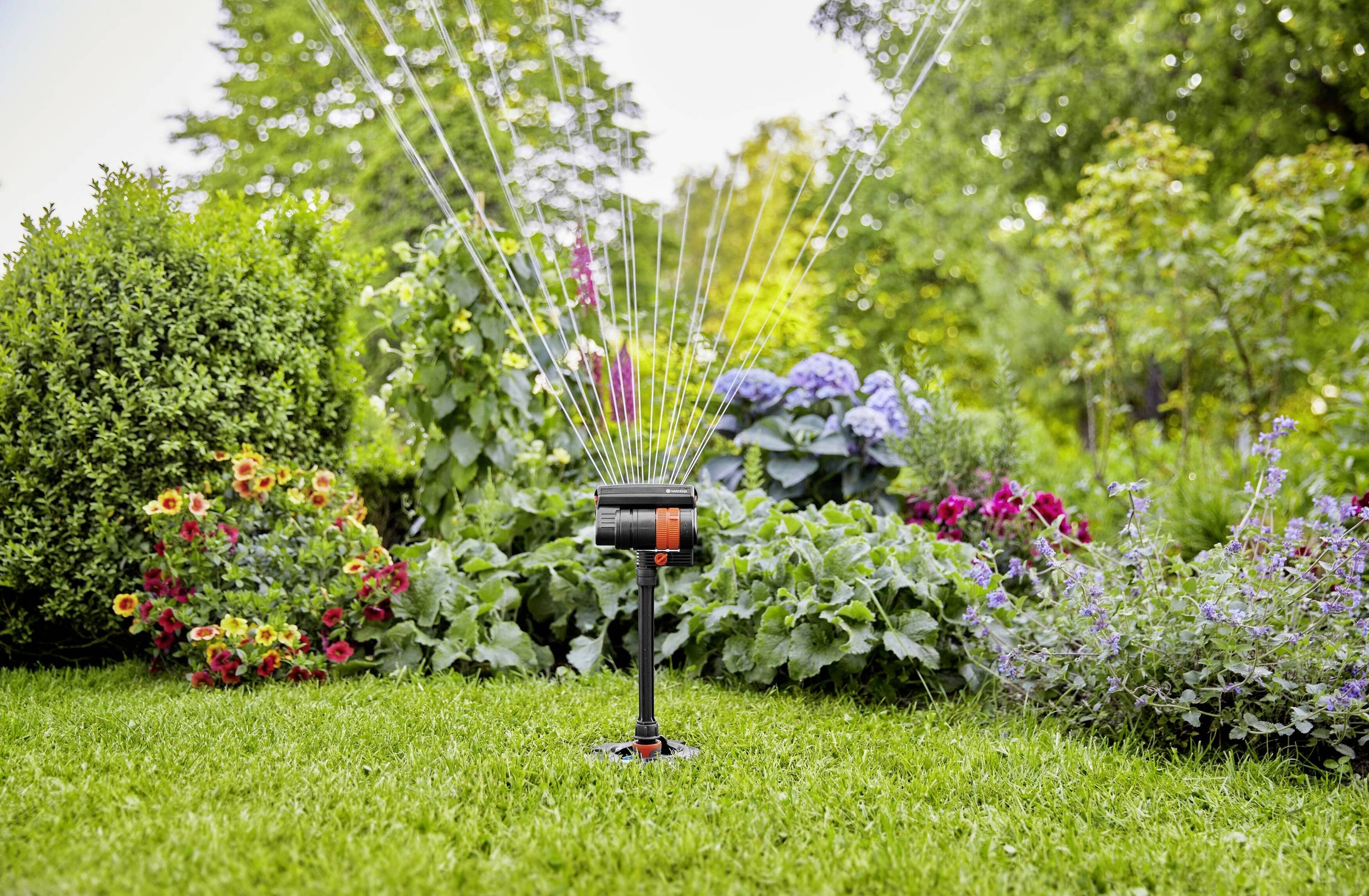 Ein Rasensprenger bewässert einen grünen Garten, umgeben von verschiedenen bunten Blumen und Sträuchern an einem sonnigen Tag.