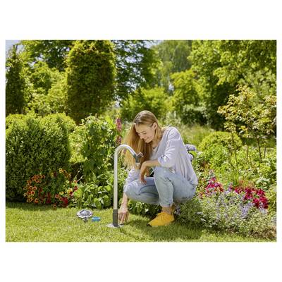 Eine Frau kniet auf dem Gras in einem Garten und dreht einen Wasserhahn auf, um Pflanzen zu bewässern, die von lebendigen Blumen und üppigem Grün unter einem sonnigen Himmel umgeben sind.