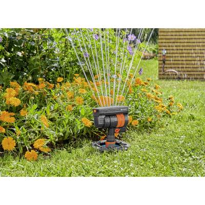 Ein Rasensprinkler bewässert einen blühenden Garten mit orangefarbenen Blumen, Wasserstrahlen fließen in einem Bogen auf den Rasen.