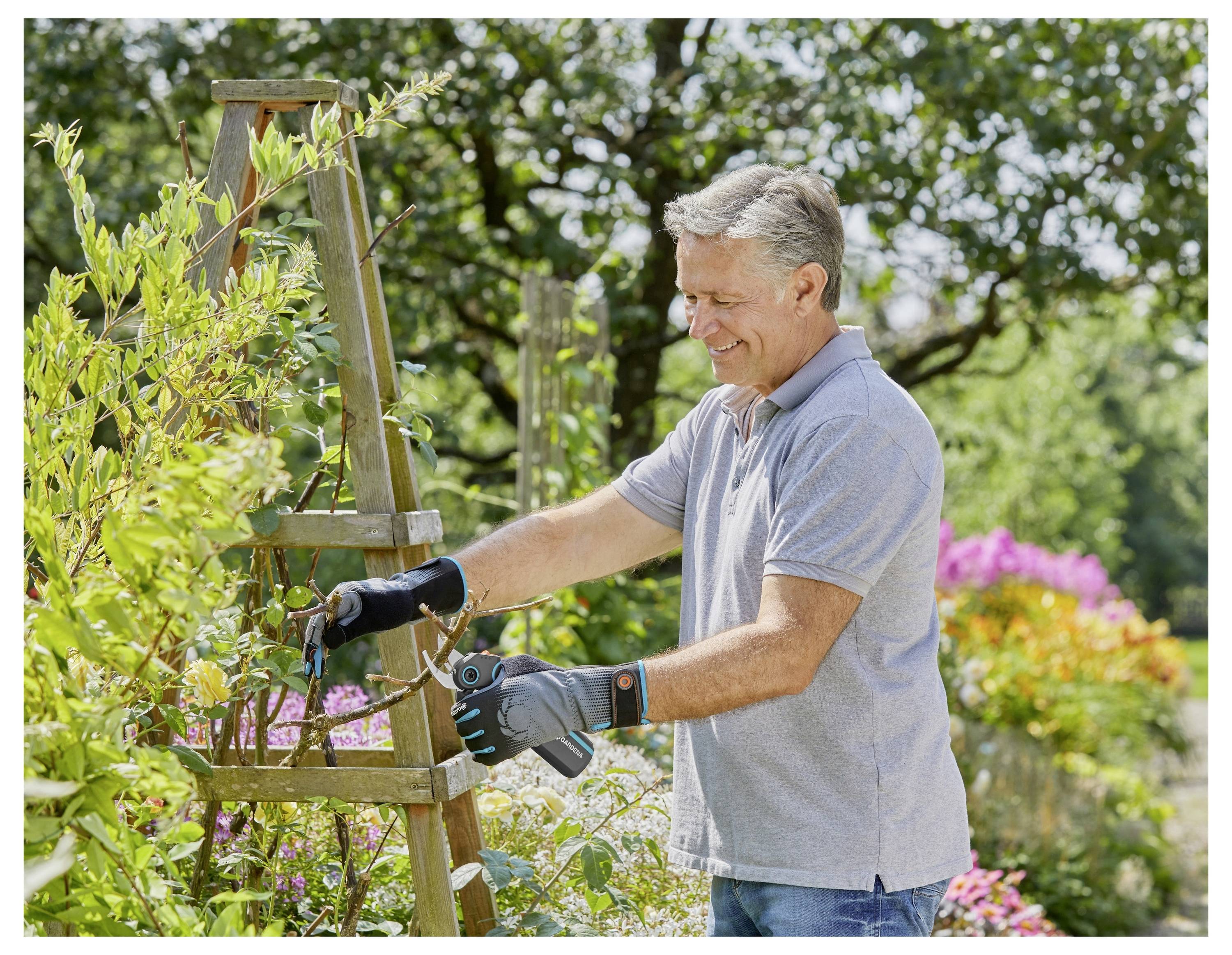 Eine Person mit Gartenhandschuhen schneidet eine Pflanze mit einer Schere zurecht und steht in einem lebendigen Garten mit blühenden Blumen und üppigem Grün.