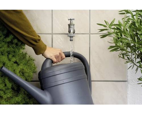 Eine Person füllt eine Gießkanne mit Wasser aus einem Wasserhahn neben grünen Pflanzen.