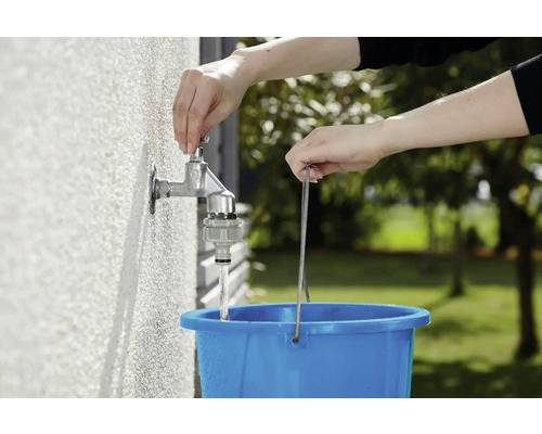 Eine Person füllt einen blauen Eimer mit Wasser aus einem Außenwasserhahn. Im Hintergrund sind Bäume zu sehen.