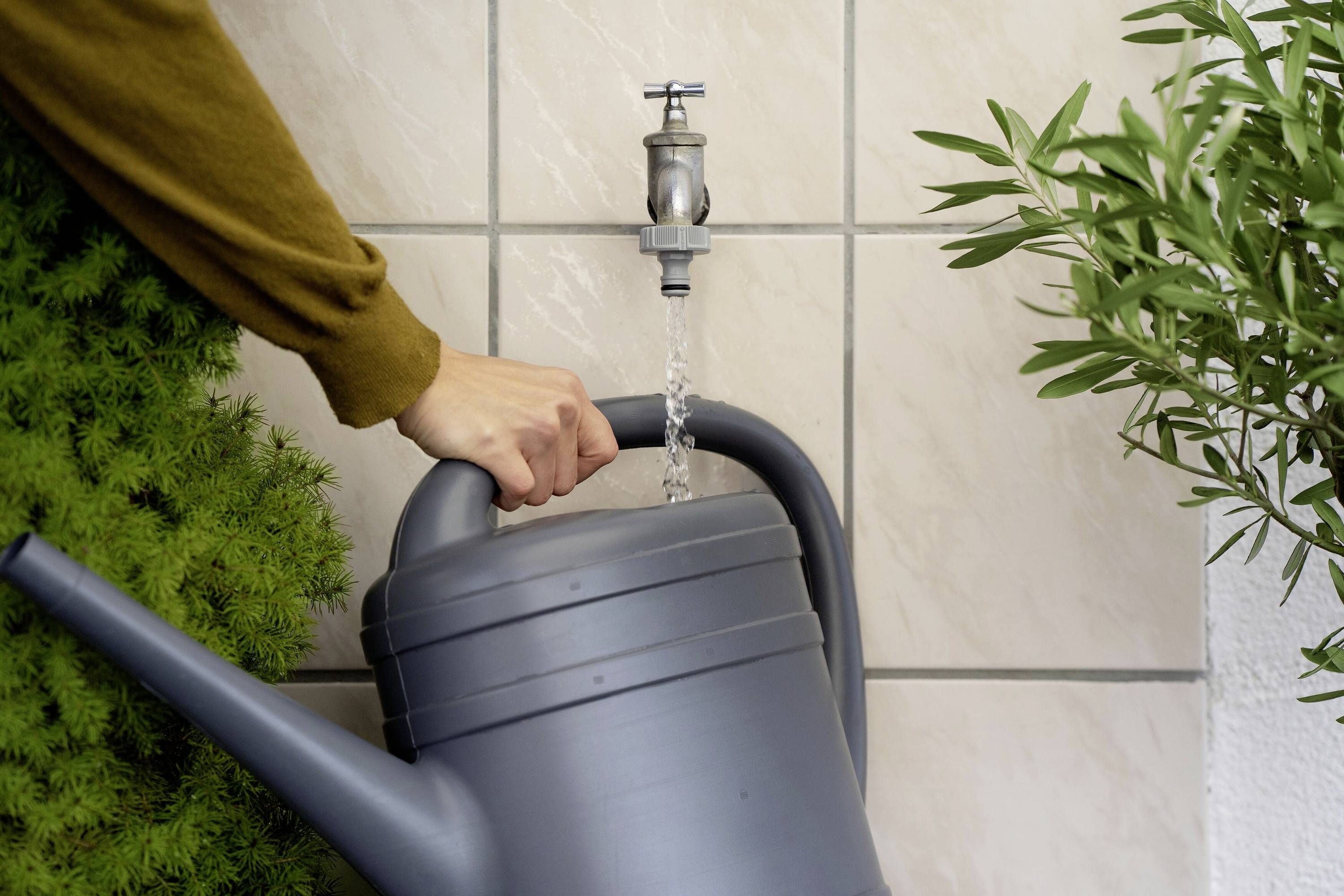 Eine Person füllt eine graue Gießkanne mit Wasser an einem Wasserhahn, umgeben von Pflanzen.