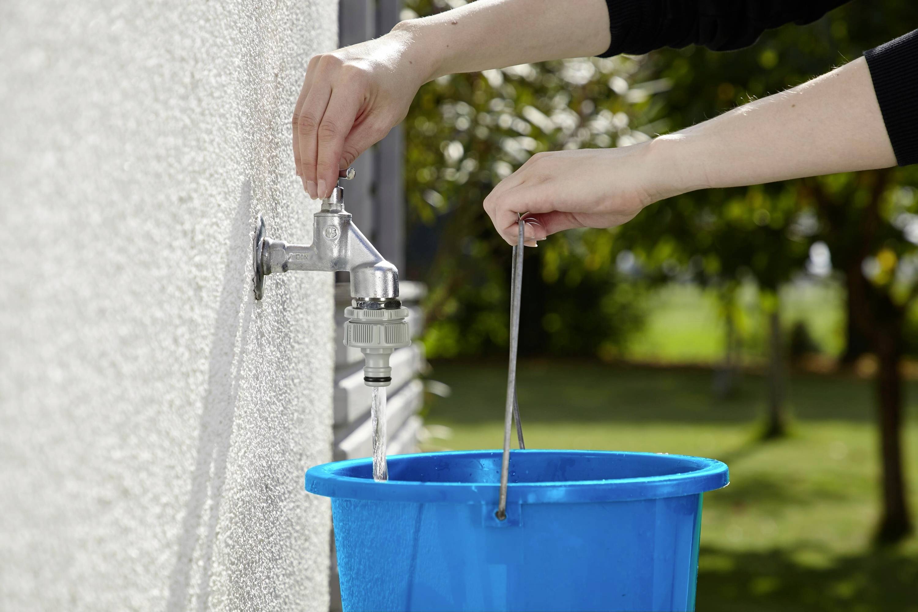 Eine Person füllt einen blauen Eimer mit Wasser aus einem Außenwasserhahn. Hintergrund zeigt grüne Bäume und Rasen.