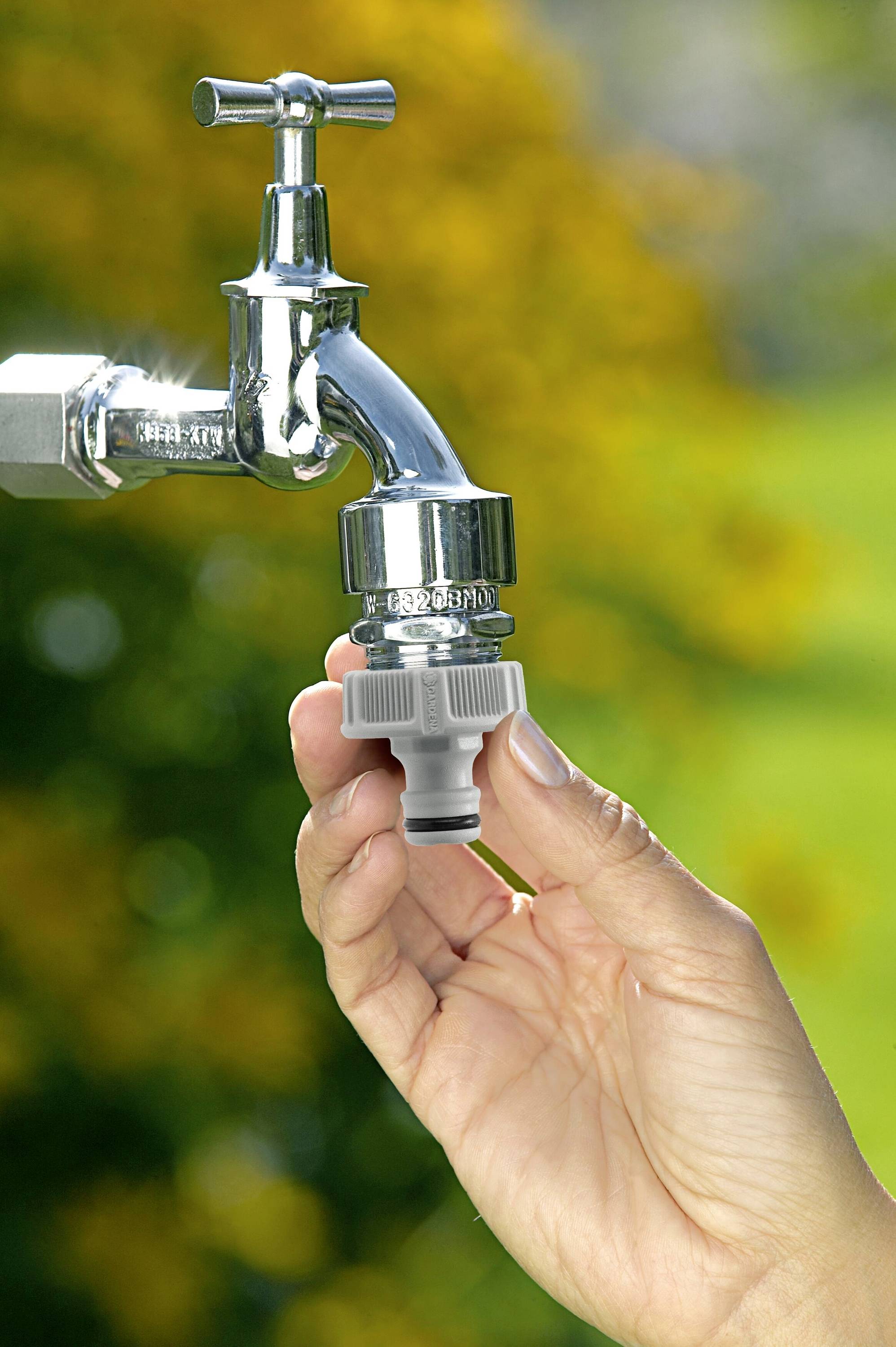 Eine Hand montiert einen grauen Schlauchadapter an einen glänzenden Wasserhahn vor einem unscharfen, grünen Hintergrund.