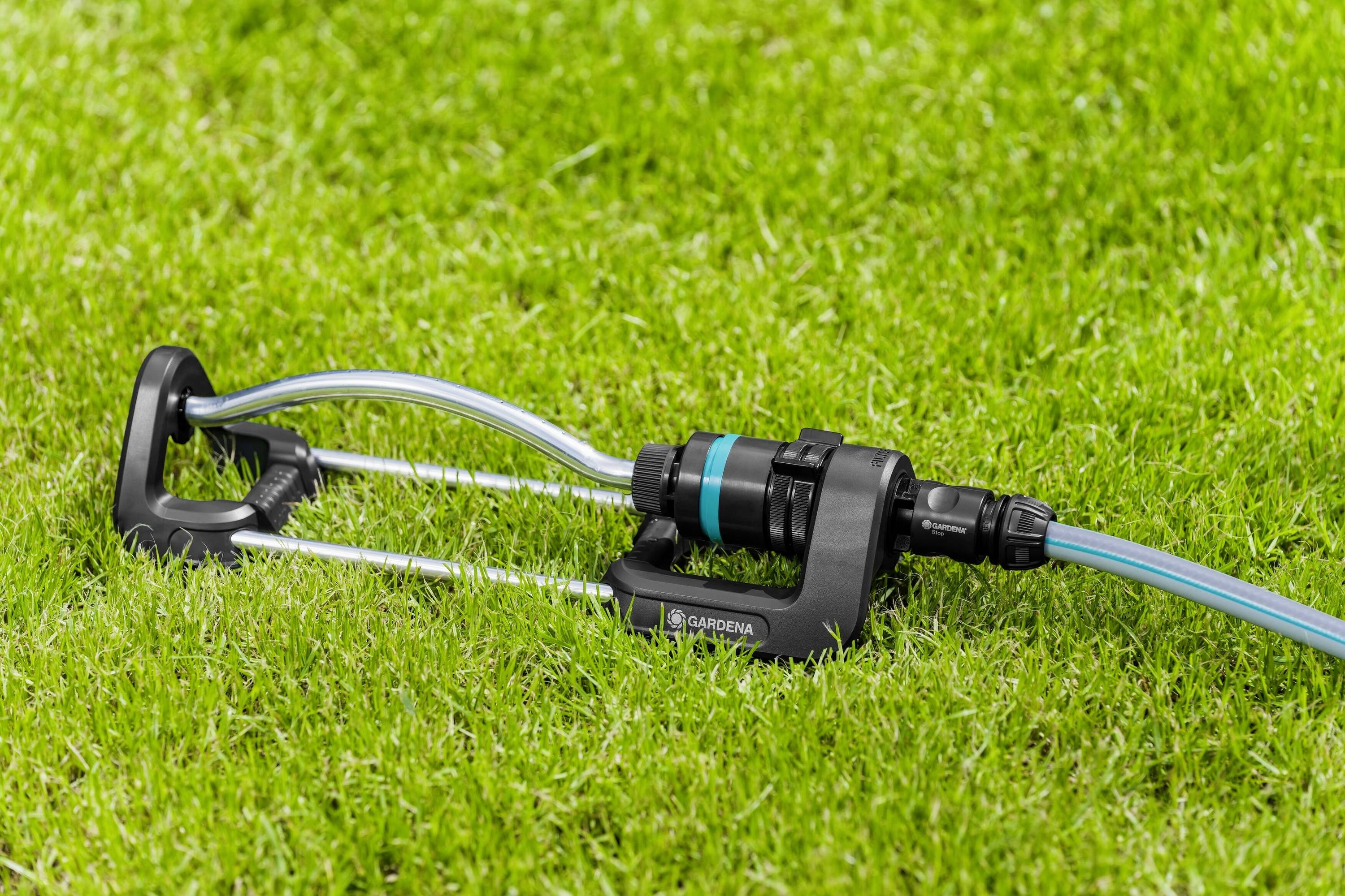 Ein Rasensprenger auf grünem Rasen, mit zwei Metallarmen und einem blauen Schlauch. Der Sprenger verteilt Wasser gleichmäßig.