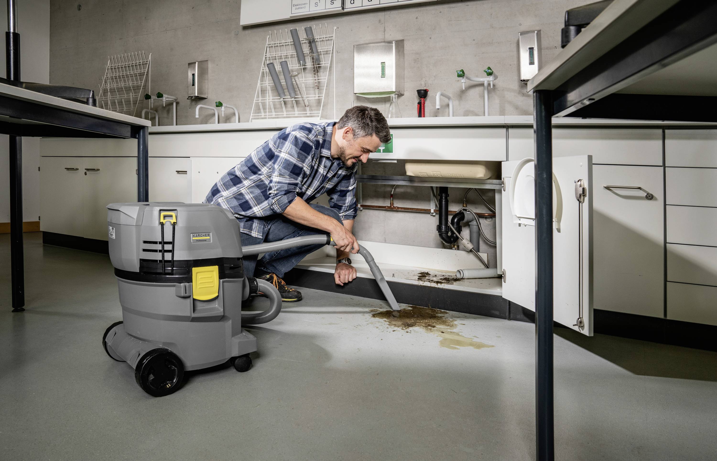 Ein Mann reinigt mit einem Industriesauger einen Abfluss in einer Werkstatt oder einem Labor. Schmutz ist sichtbar auf dem Boden.
