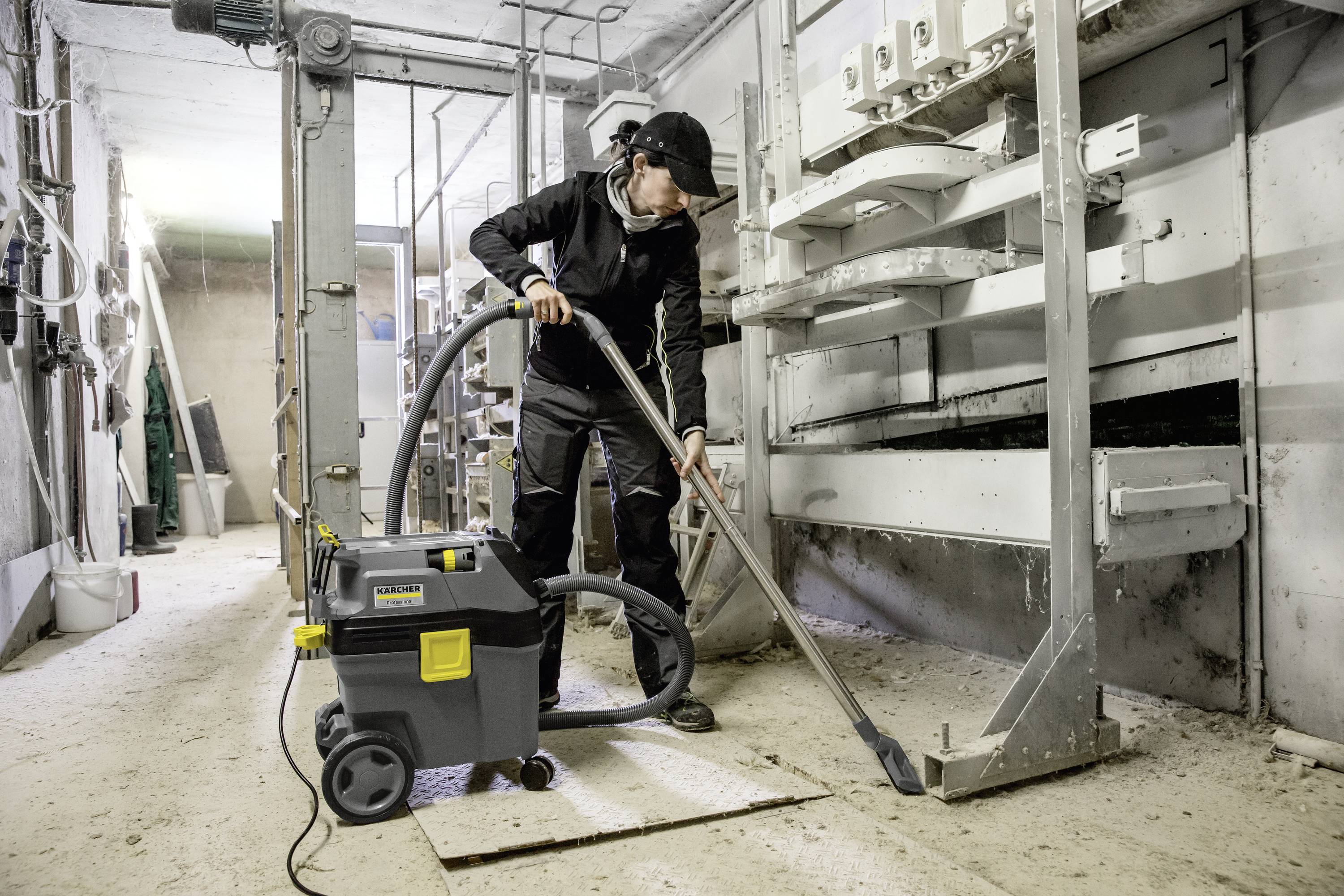 Eine Person reinigt mit einem Industriestaubsauger einen großen, staubigen Raum. Im Hintergrund sind Maschinen und Rohre zu sehen.