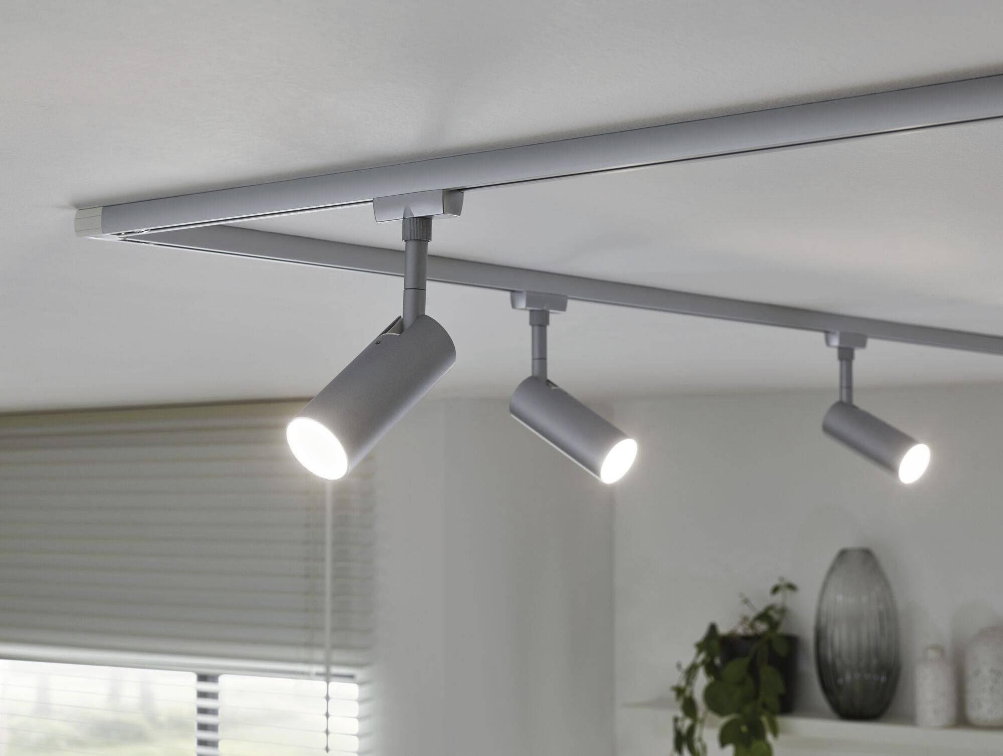 Moderne Deckenstrahler mit drei verstellbaren Leuchten in einem Wohnraum. Links sind Fenster mit Jalousien und rechts Dekorationen sichtbar.